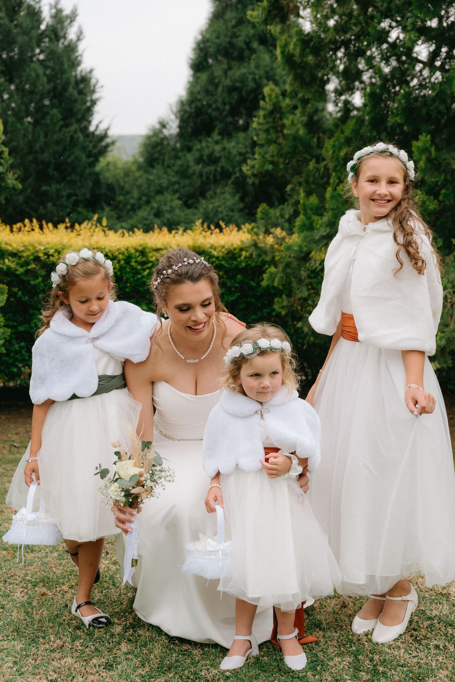 Young flower girls in white dresses playing and posing together outdoors at a wedding.