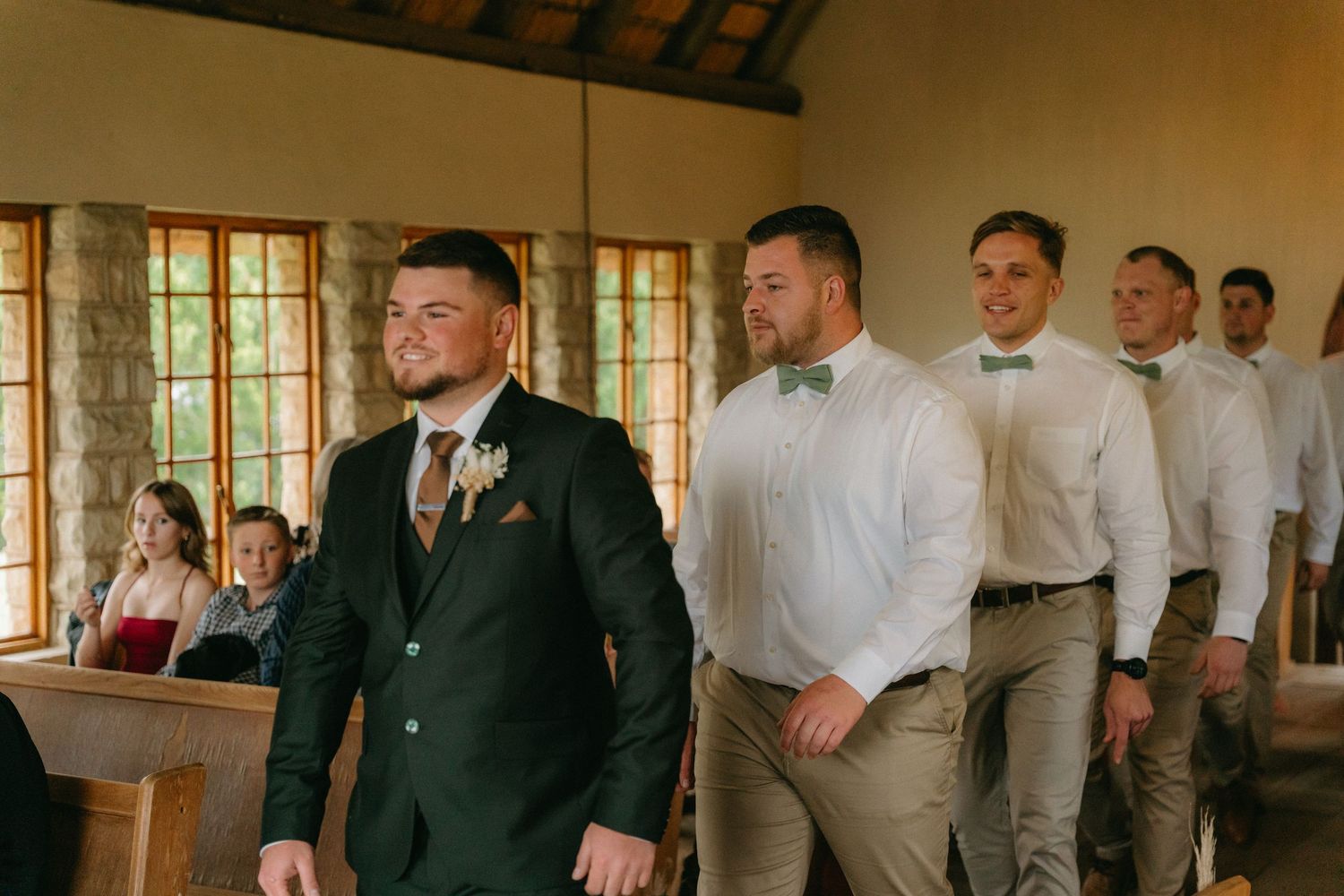 Wedding party members gather in a rustic indoor venue with wooden beams and windows.