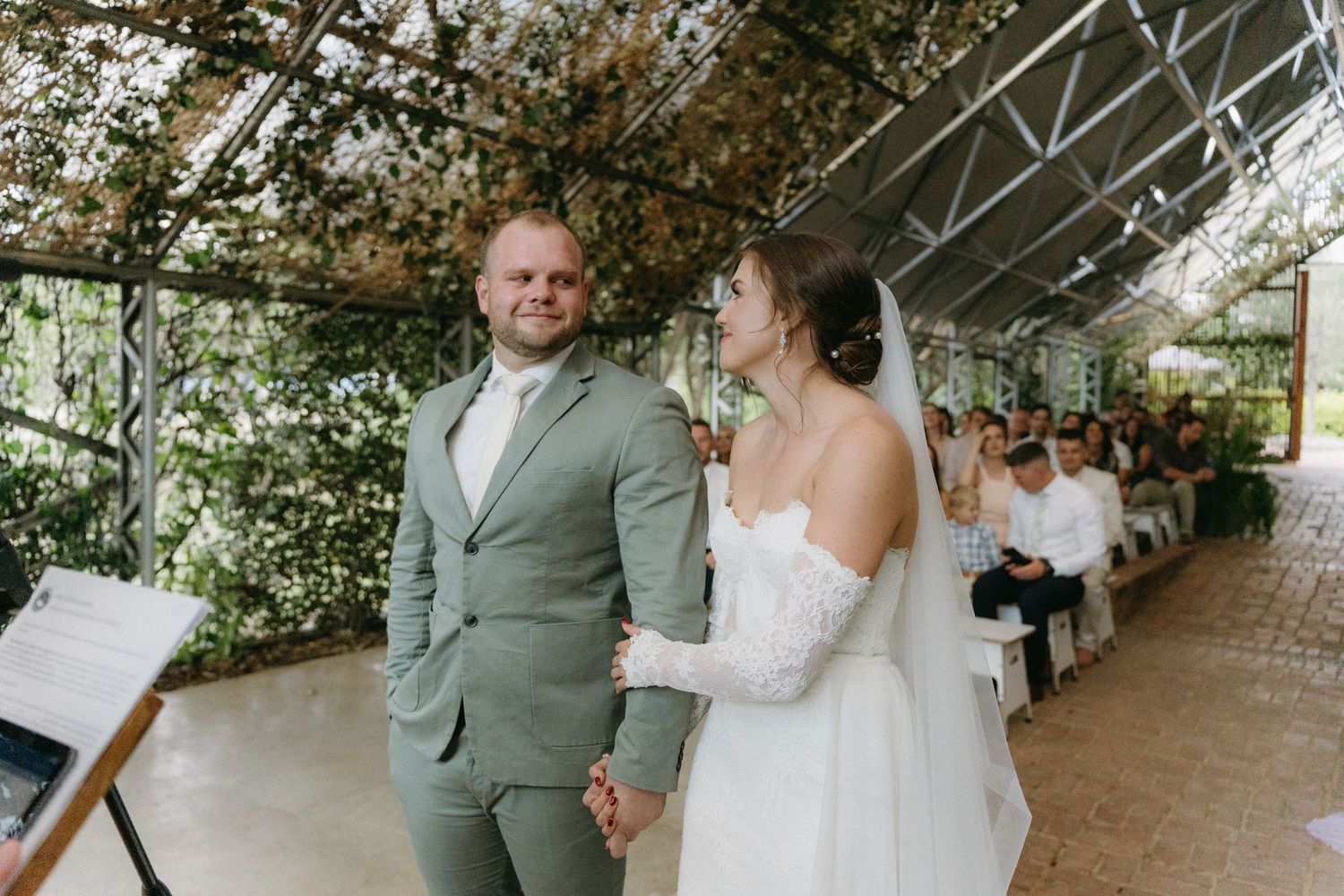 A wedding ceremony under a vine-covered pergola with guests seated on white chairs.