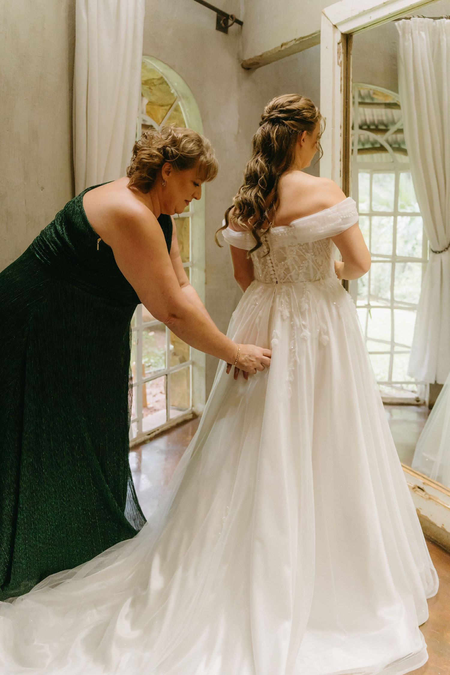 A bridesmaid in emerald green helps adjust the train of a bride's off-shoulder wedding gown.