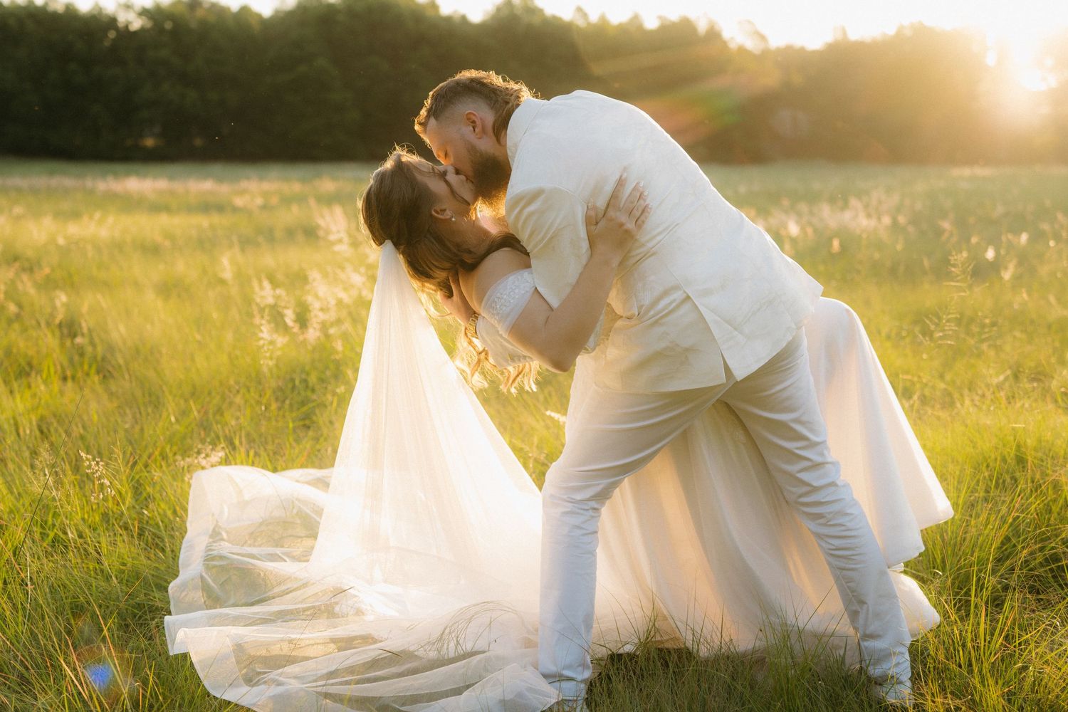 A romantic wedding couple shares an intimate dip pose in a sunlit meadow at golden hour.