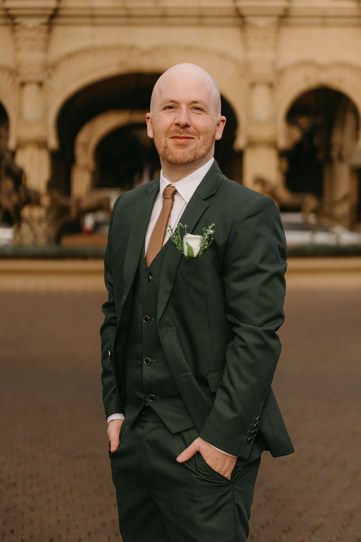Groom in dark three-piece suit with boutonniere poses against architectural arches at wedding venue.