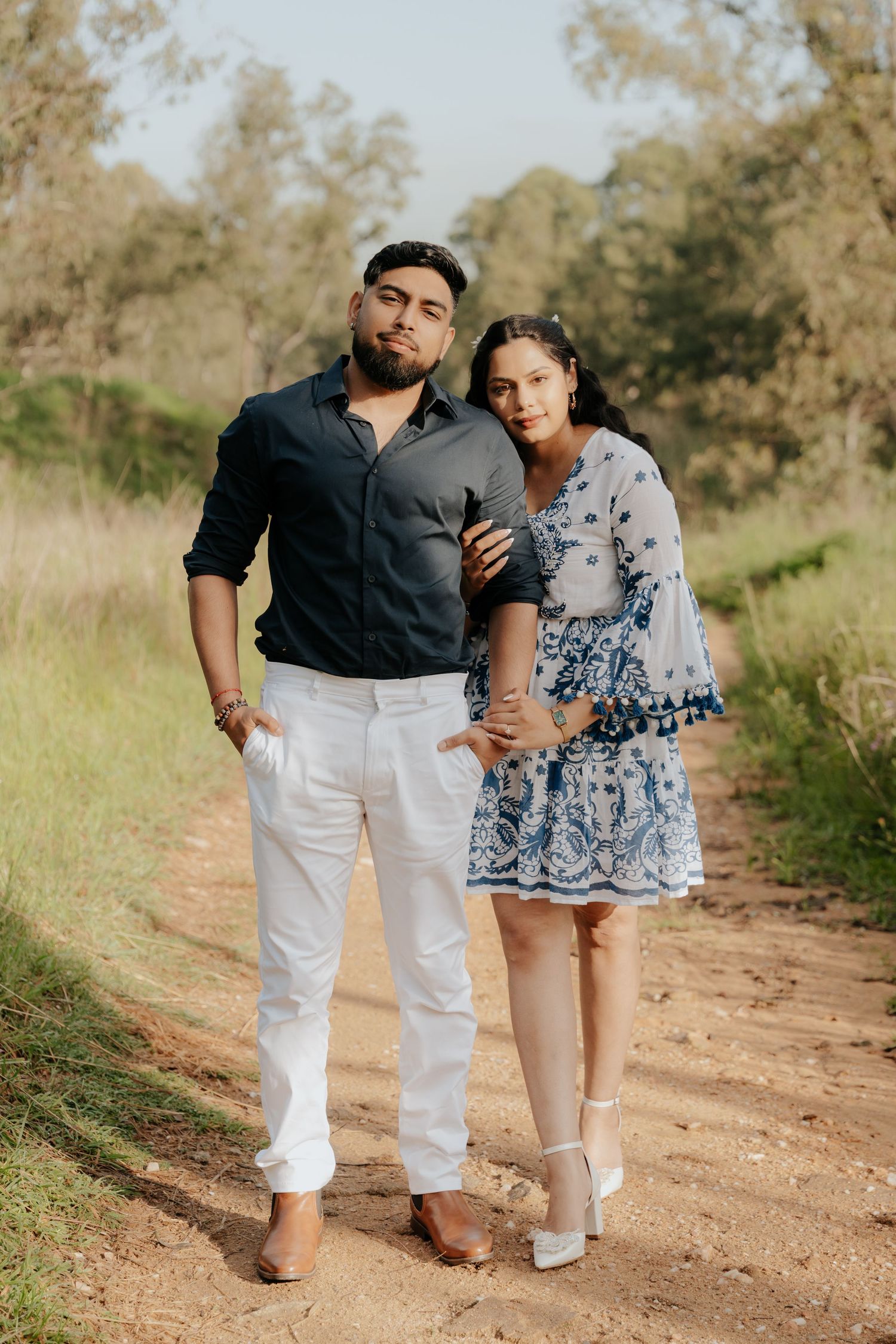 A couple in coordinated white and blue outfits walks playfully along a dirt path in a natural outdoor setting.