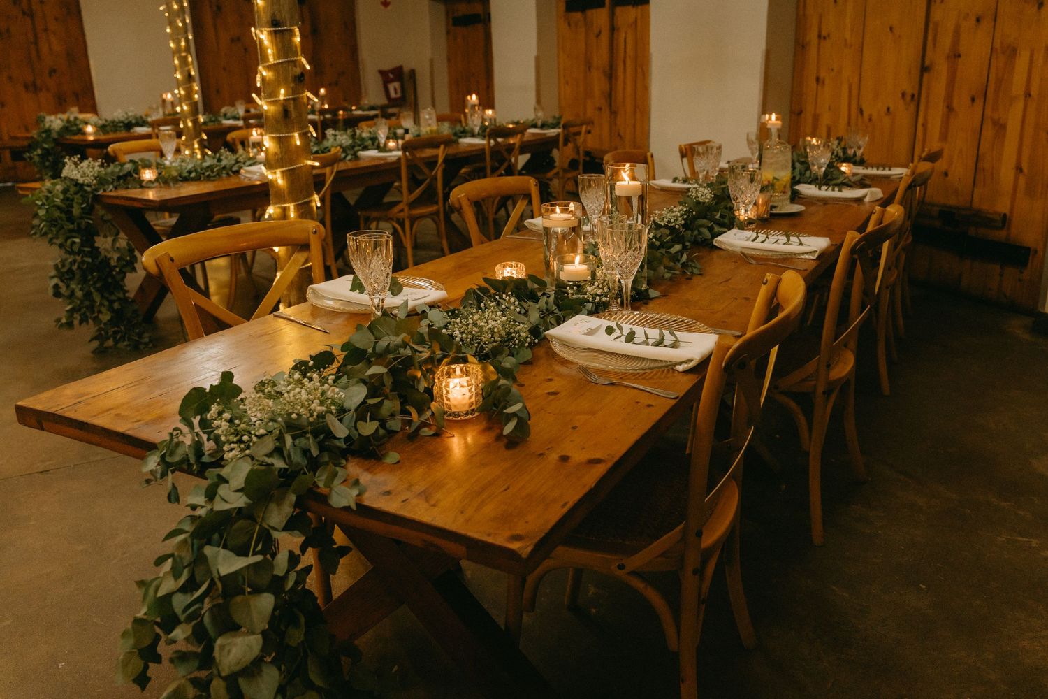 Rustic wedding reception setup with wooden tables, greenery and warm lighting.