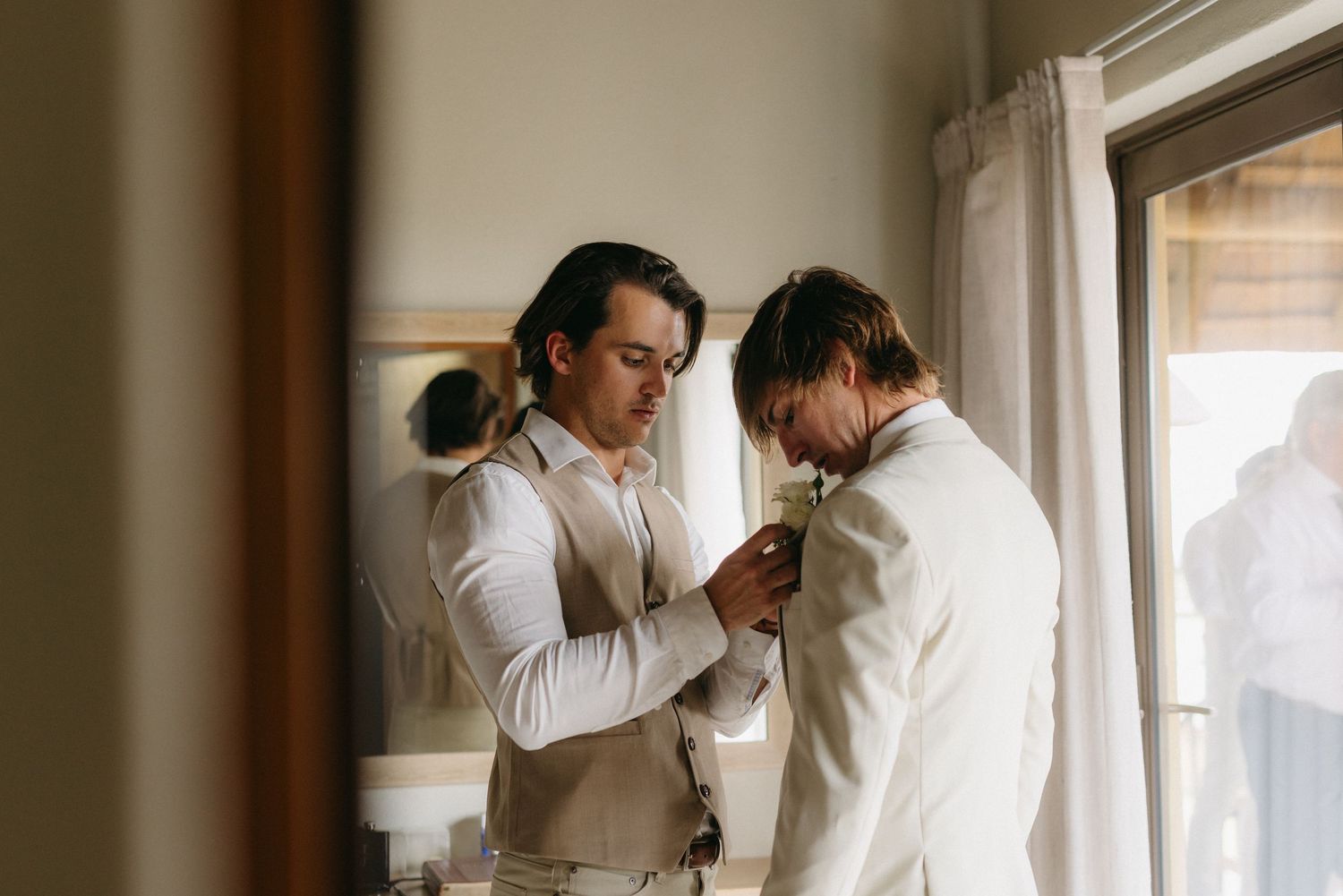 Two people help adjust formal attire near a window in preparation for a wedding.