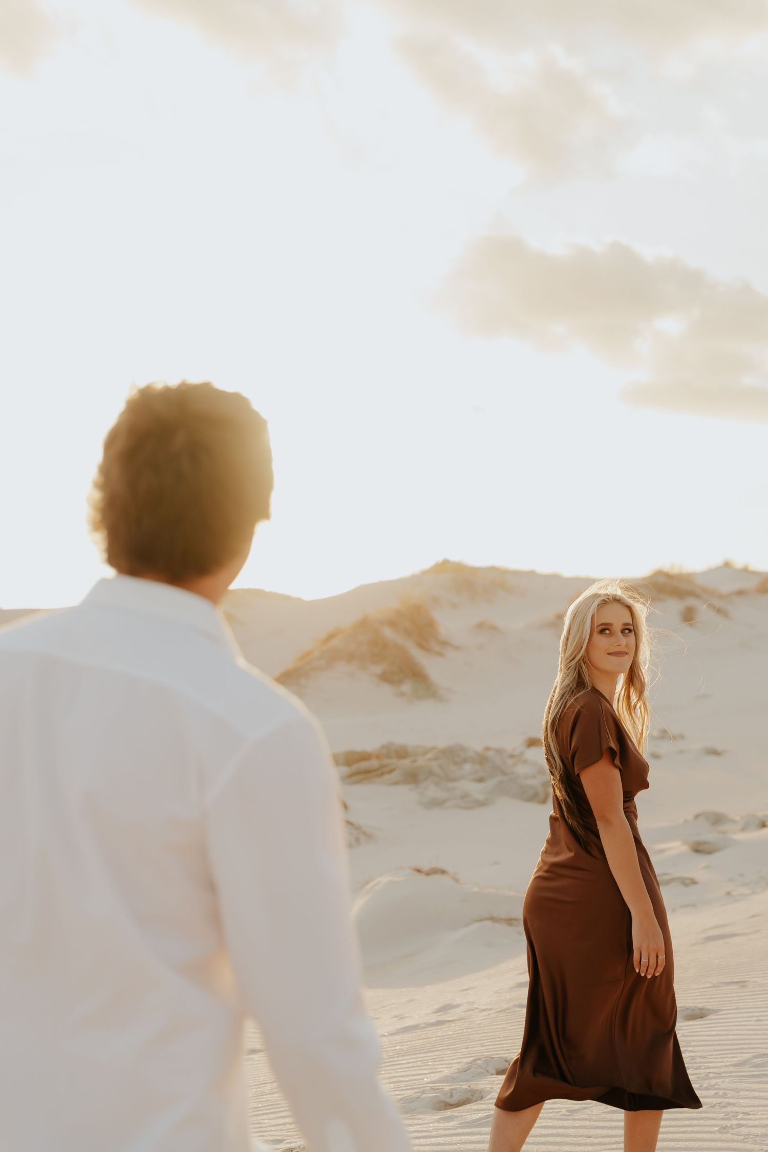 A woman in a brown dress walks on sandy dunes at golden hour, glancing back at a man in a white shirt.