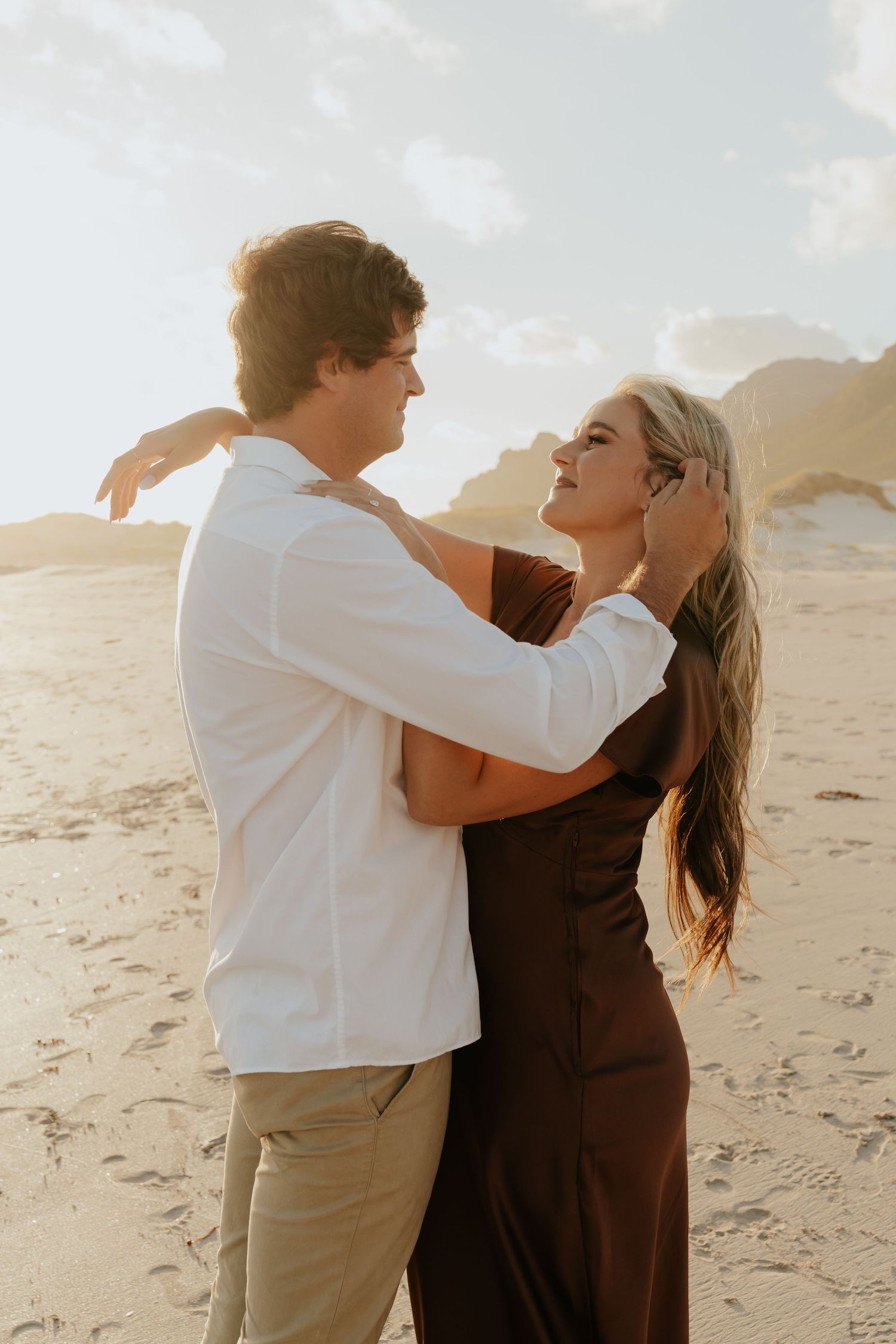 A couple embraces on a sandy beach at golden hour, the woman in a brown dress smiling up at the man in a white shirt.