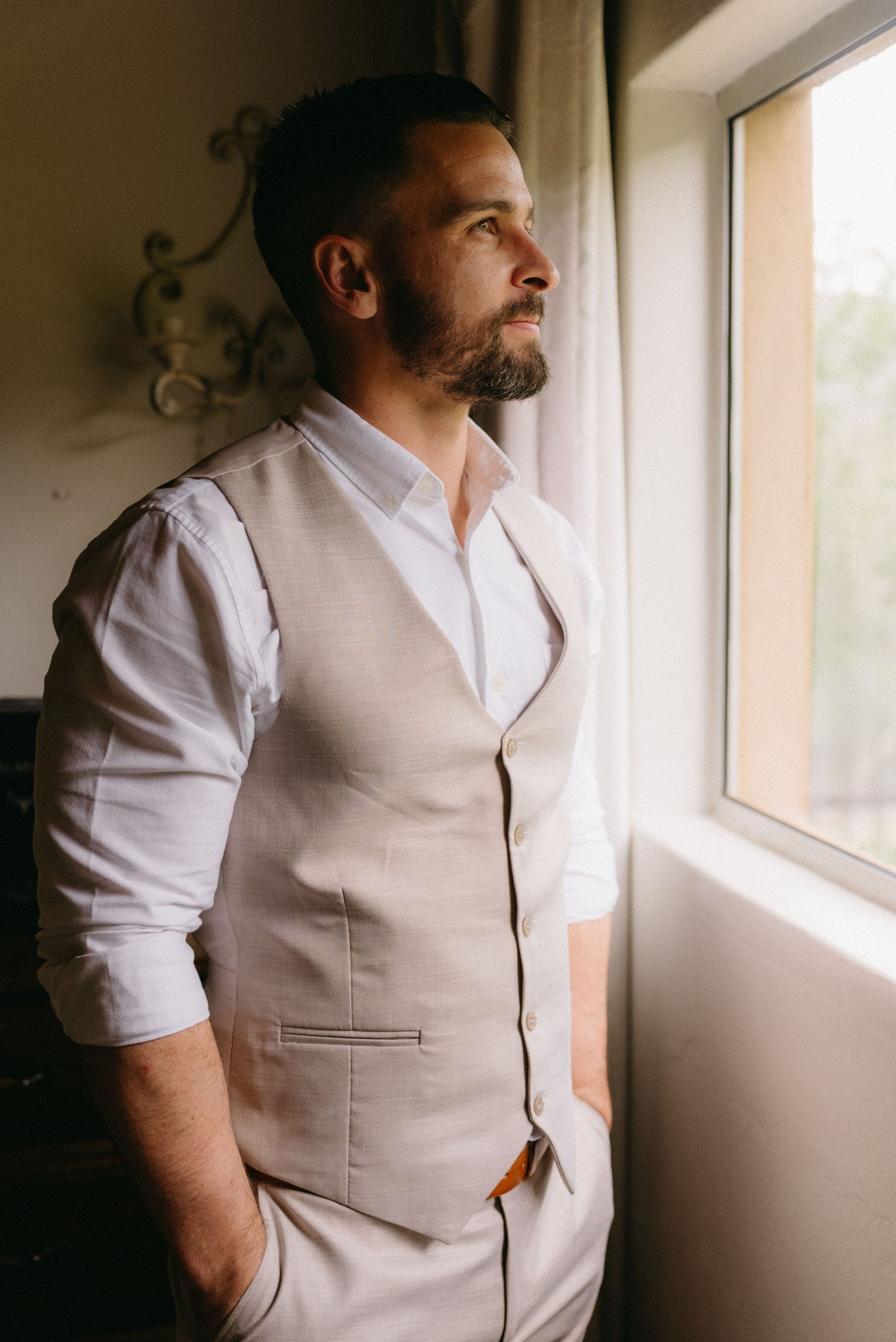 Groom wearing a light beige vest and white shirt looking towards a window in natural light.