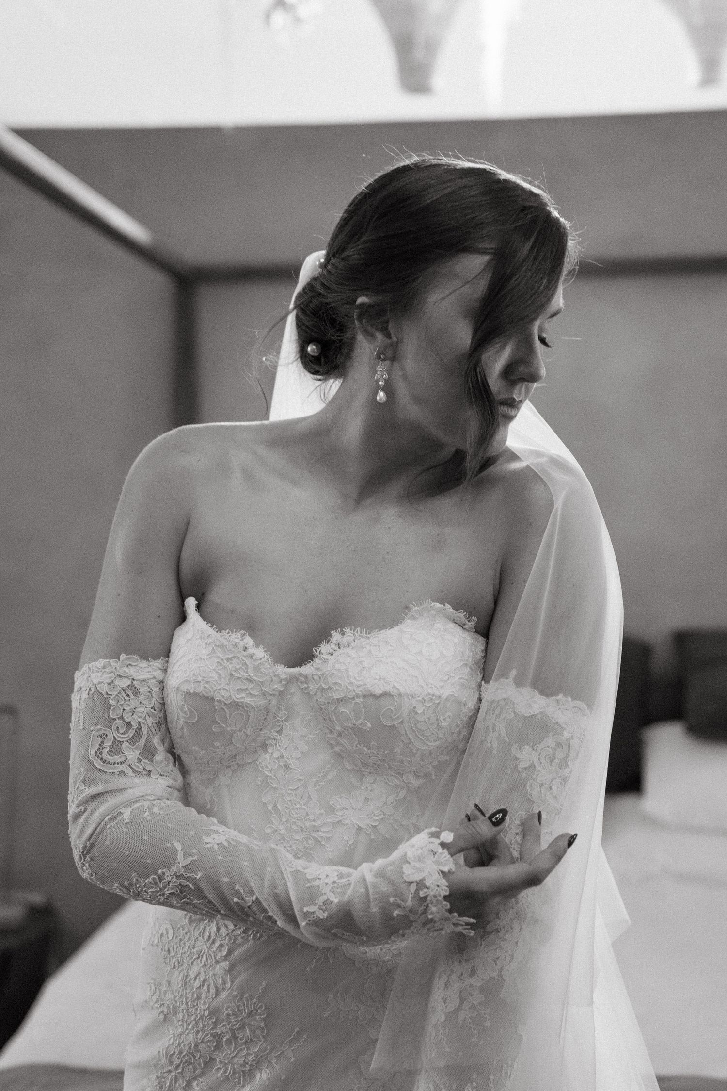A bride in an elegant lace wedding dress with long sleeves looks down while wearing a veil in a black and white photo.