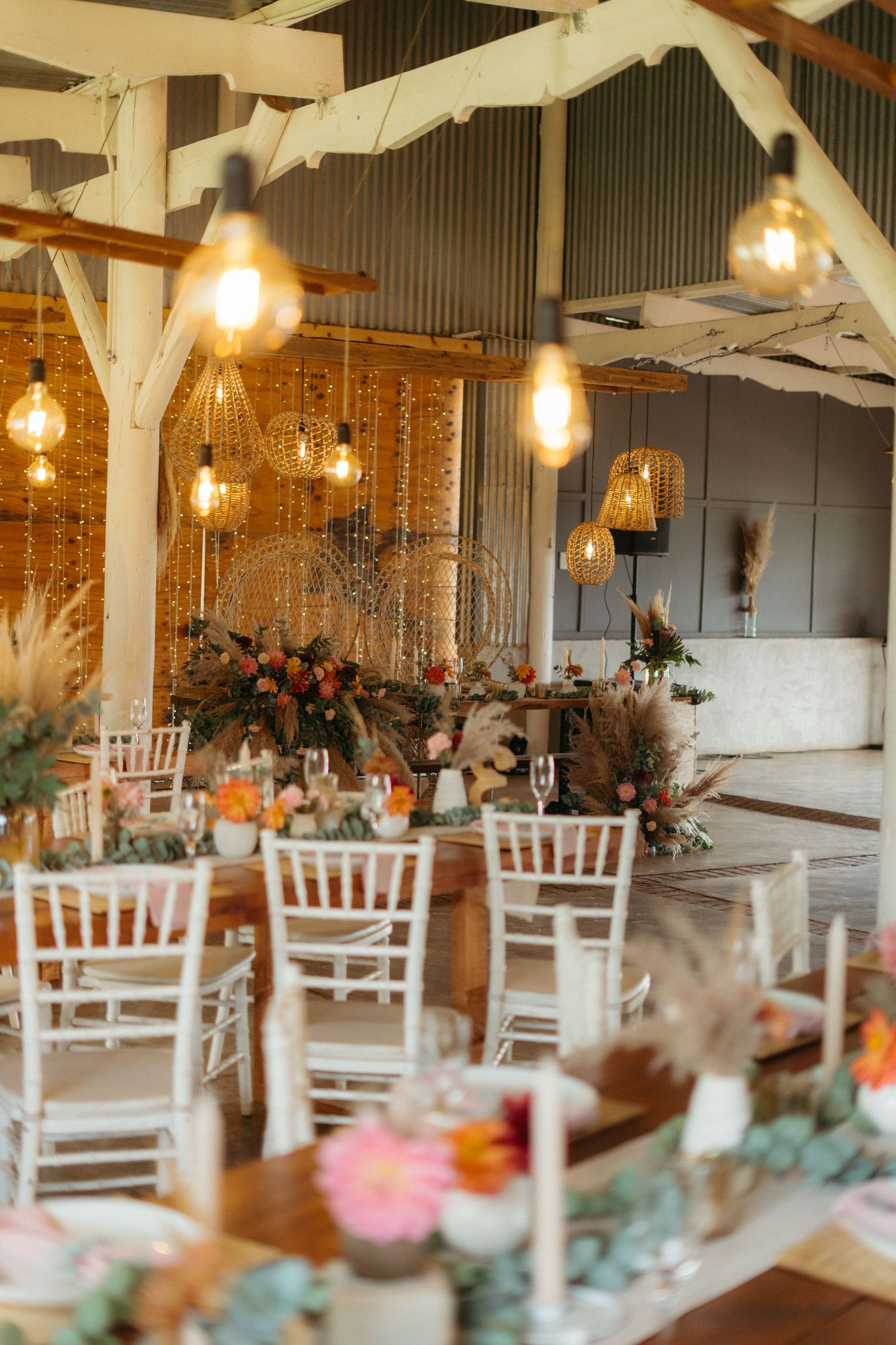 Rustic wedding reception venue with white chairs, hanging Edison bulbs, and floral arrangements on long dining tables.