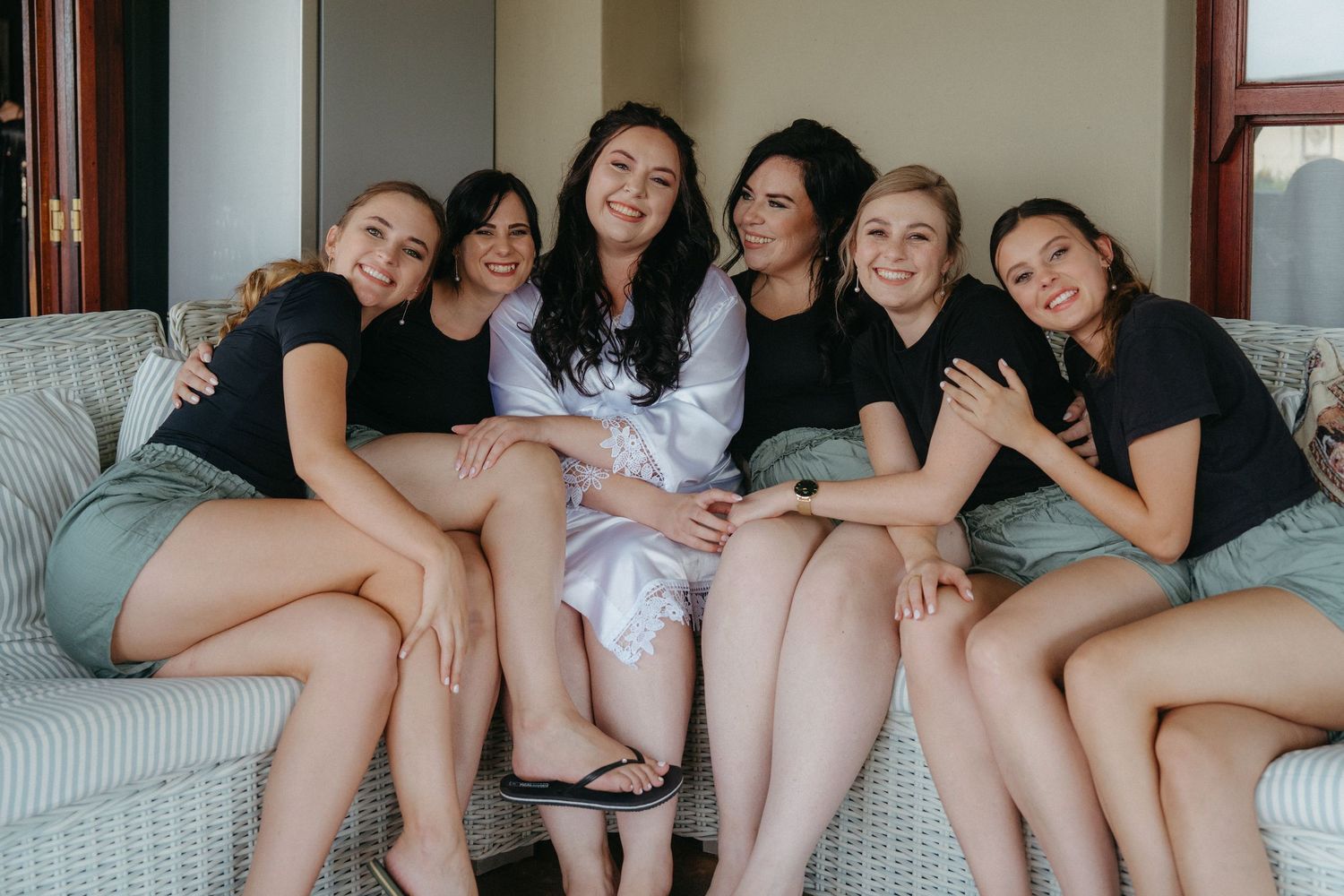 Bridesmaids in matching black tops and shorts lounging together on a light gray couch.