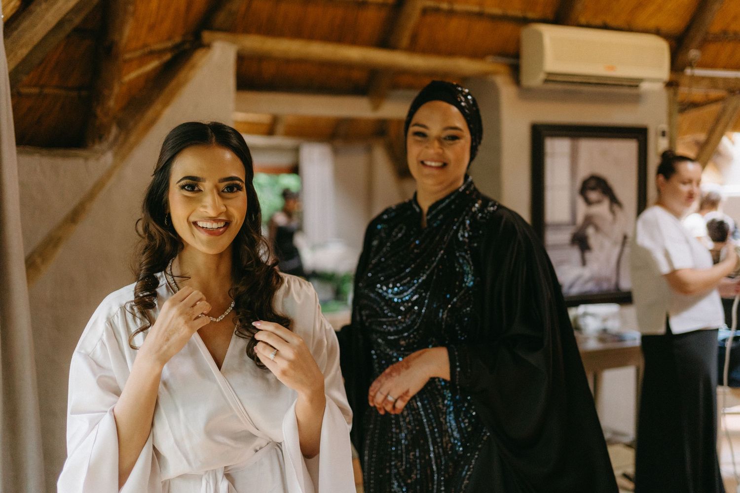 Two women getting ready in robes at rustic venue with wooden beams.
