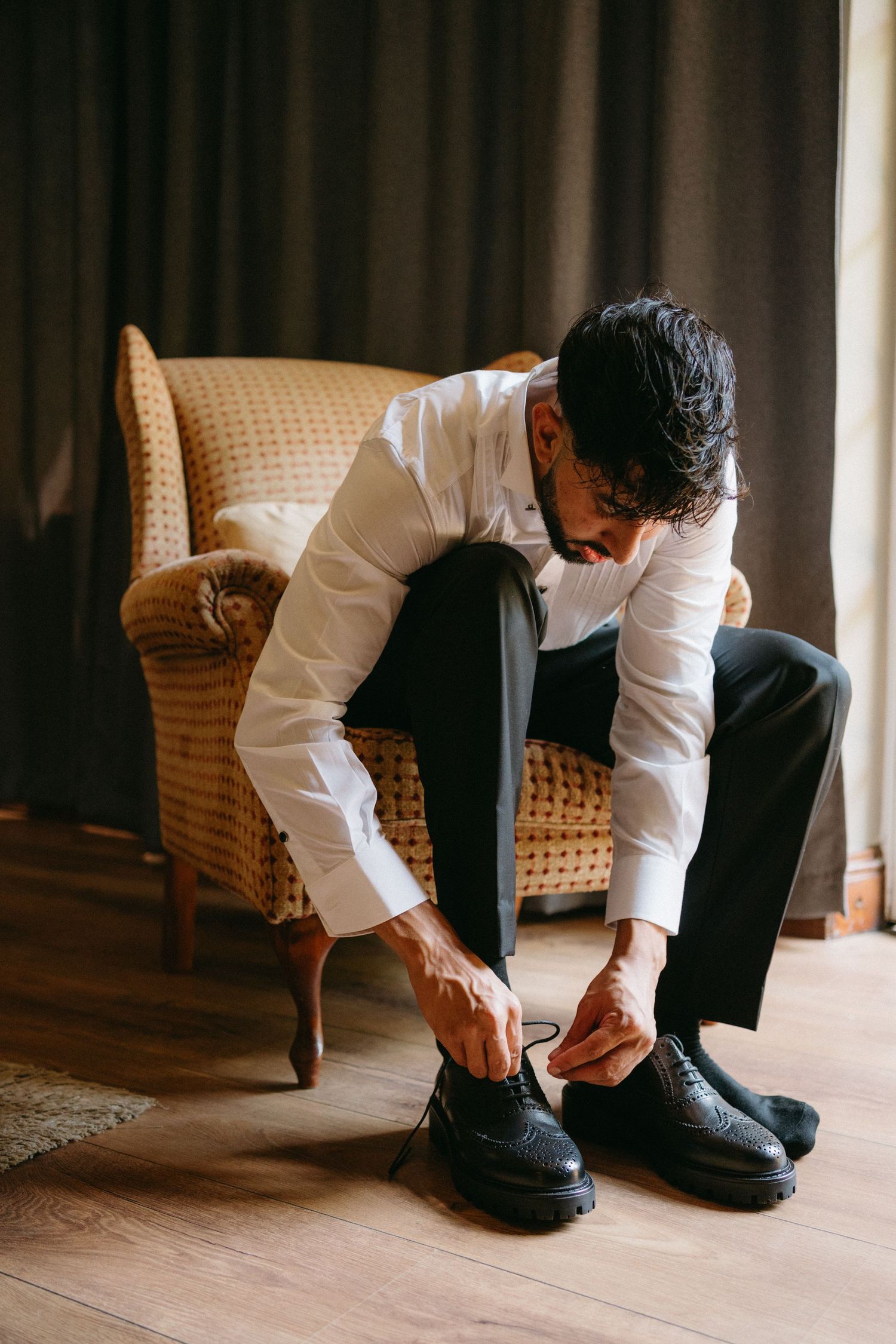 A person in formal attire sits in a woven armchair while tying the laces of black leather dress shoes.
