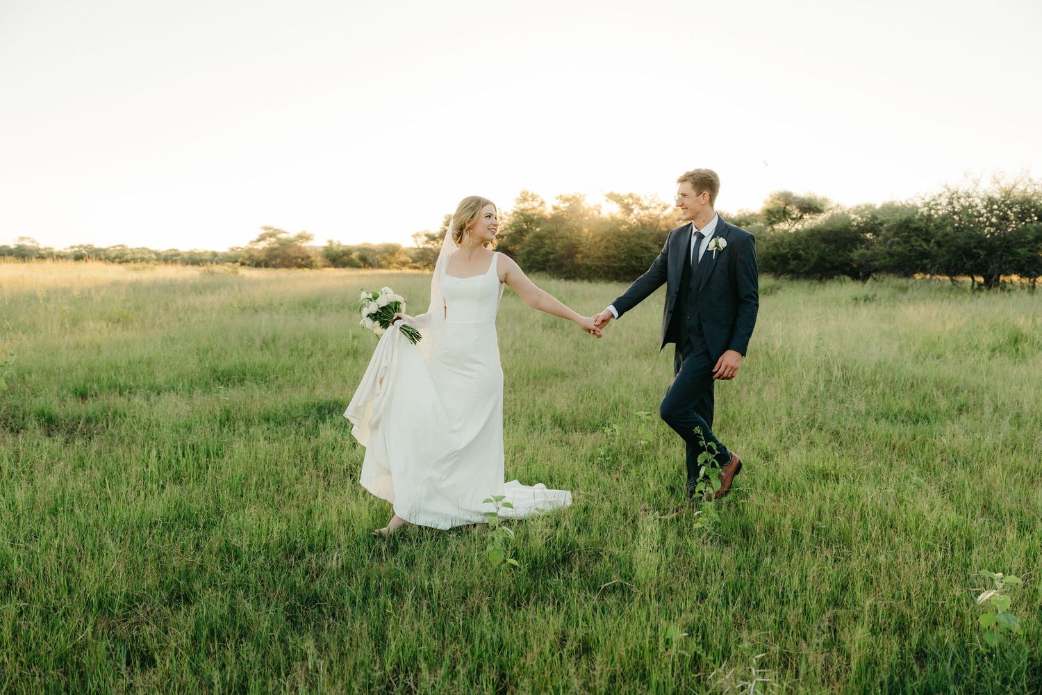 A couple walks hand in hand through a sunlit field in wedding attire during golden hour.