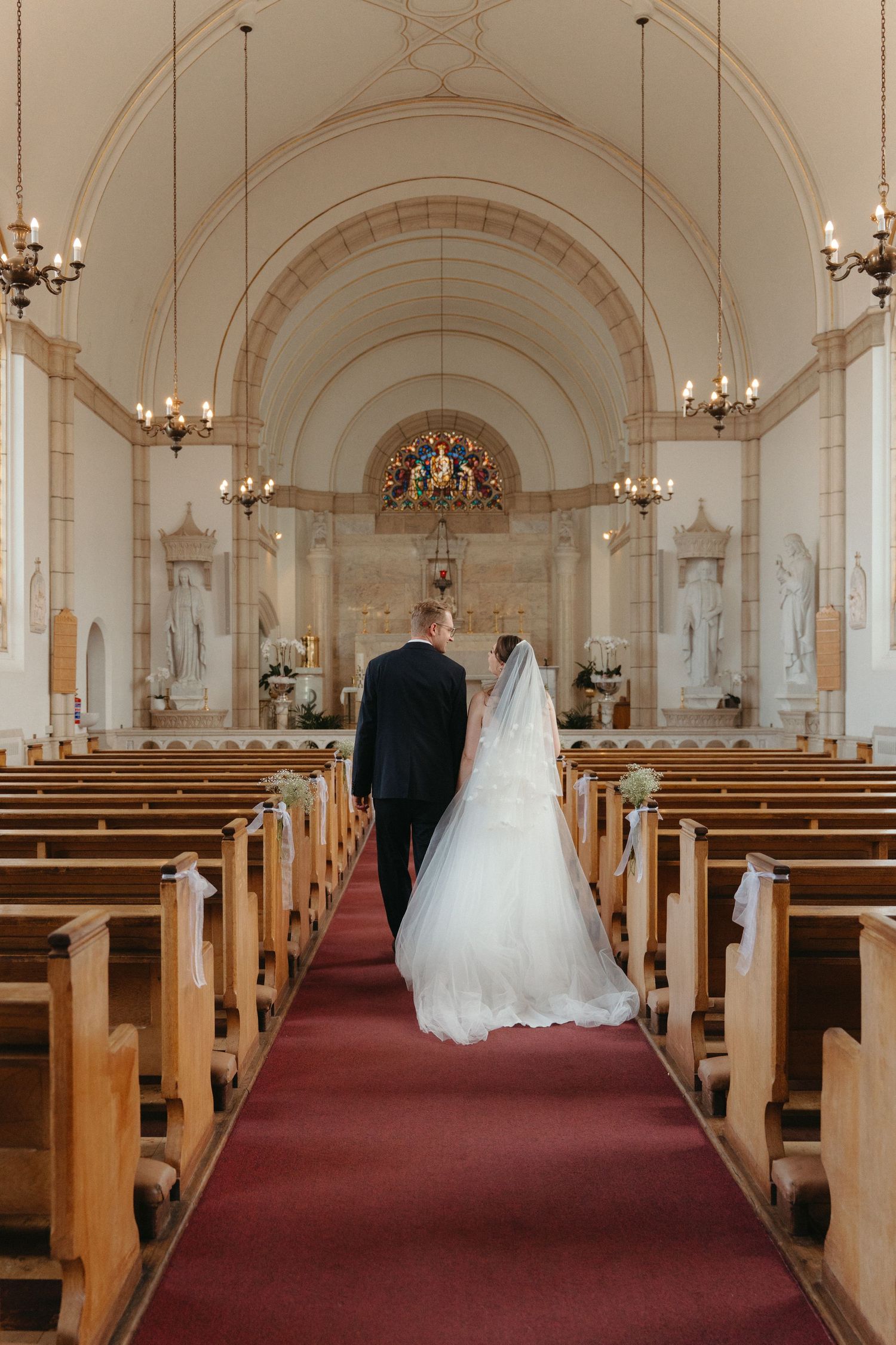 A panoramic view of a Nazareth House Chapel with wooden pews, chandeliers, and a couple in wedding attire at the altar.