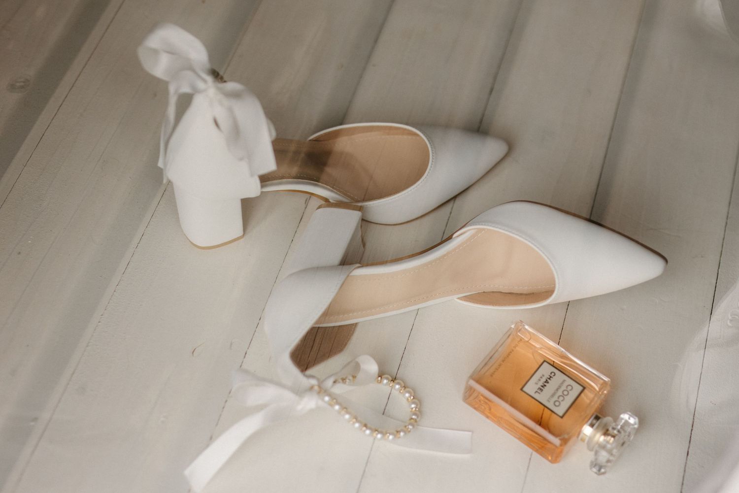White wedding heels with perfume bottle and accessories arranged on wooden surface.
