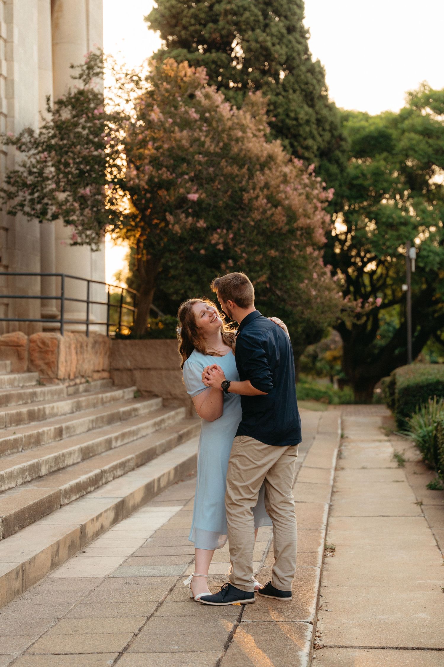 A couple embraces on stone steps at sunset with trees and greenery in the background.