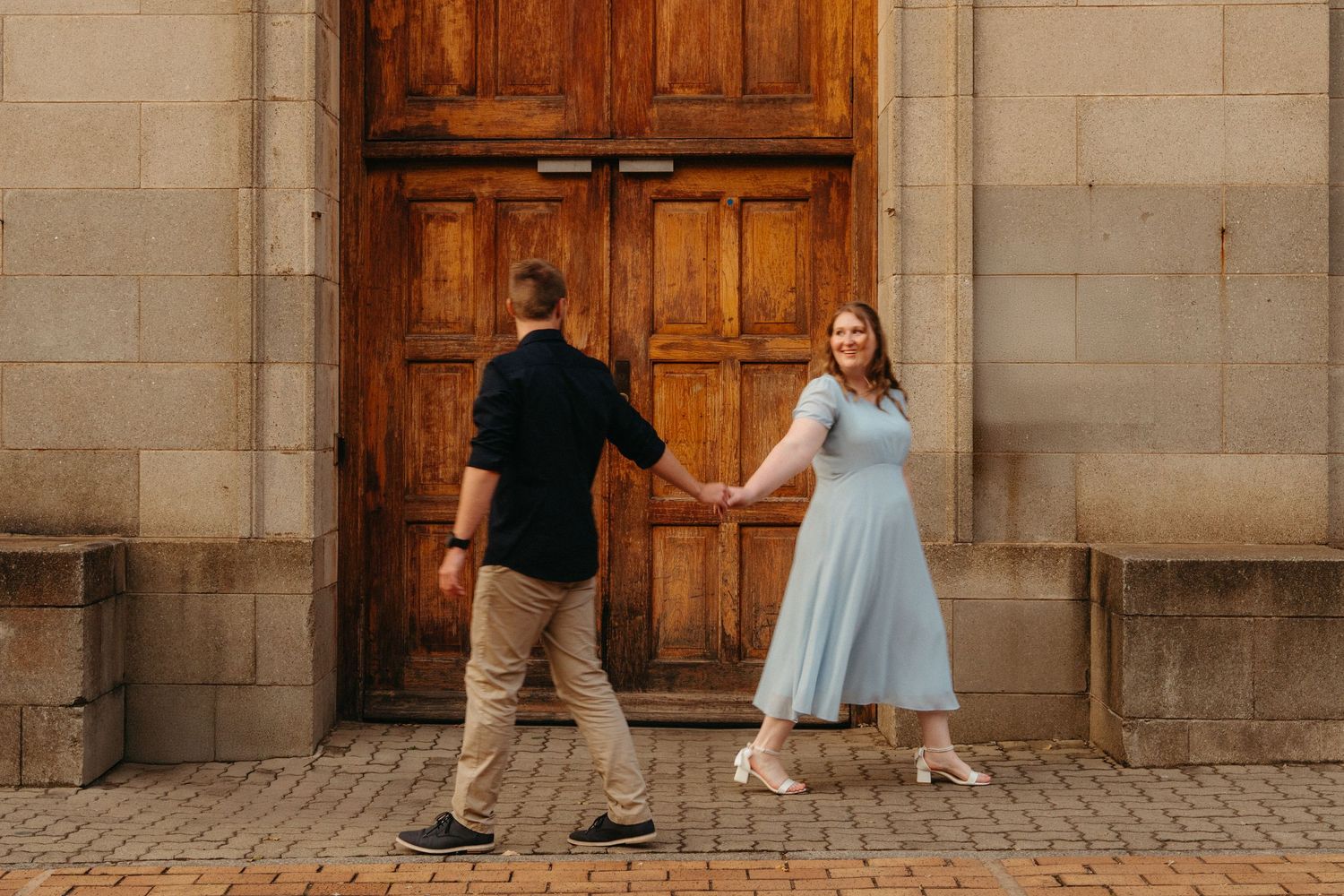 A couple in casual attire dances together on a stone patio in front of ornate wooden doors.
