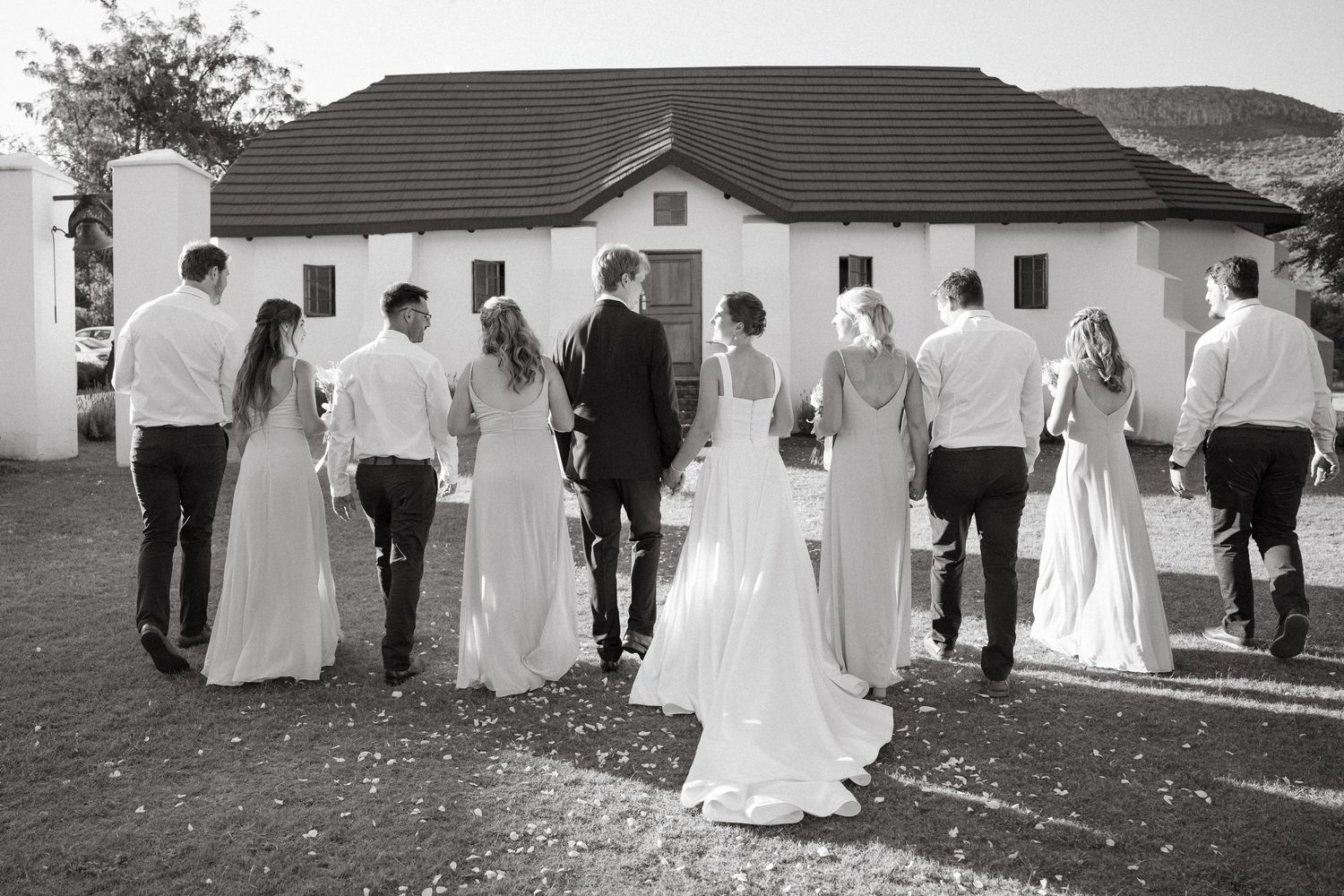A wedding party walks in a line together outside a white building in black and white photography.