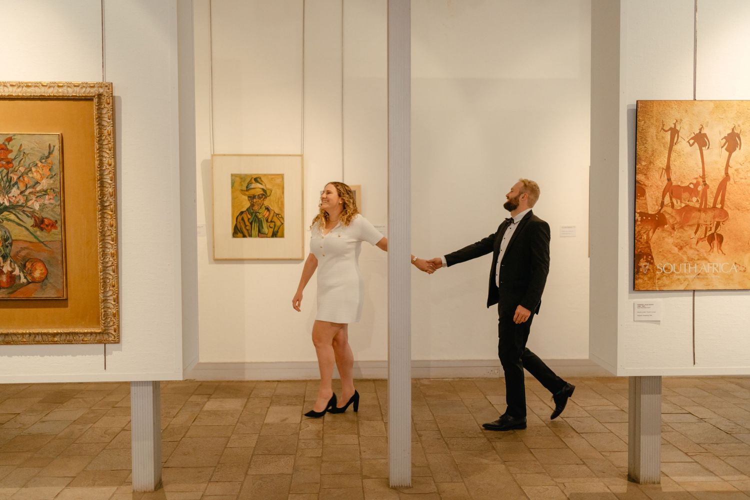 Playful couple dancing in an art gallery surrounded by framed paintings on white walls.