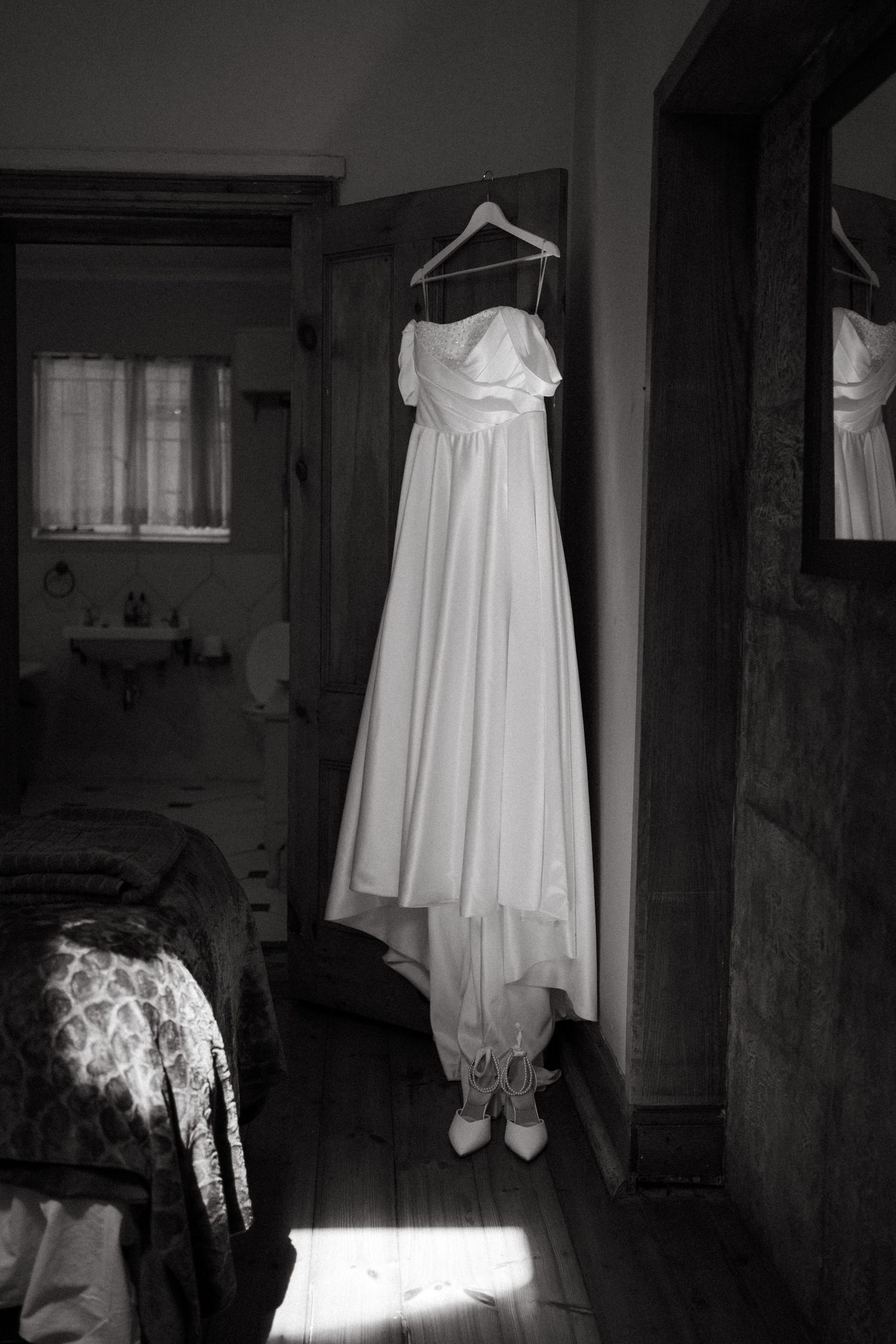 A white wedding dress hangs on a hanger in a dimly lit room with dramatic lighting and shadows.