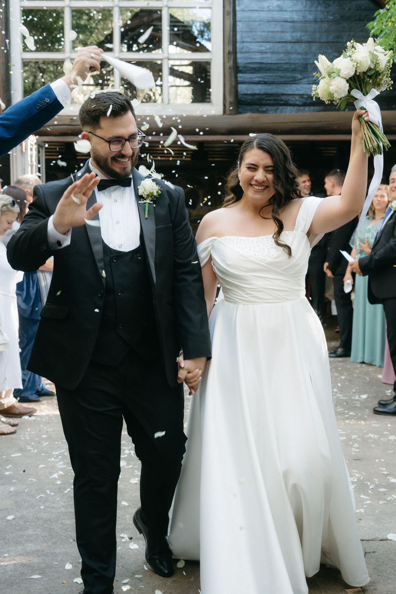 A newlywed couple celebrates with joy as wedding guests shower them with confetti outside a blue building.