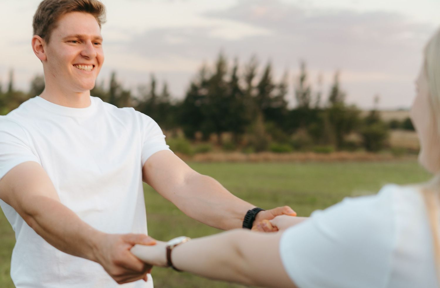 Two people in white shirts holding hands in a grassy field at sunset.