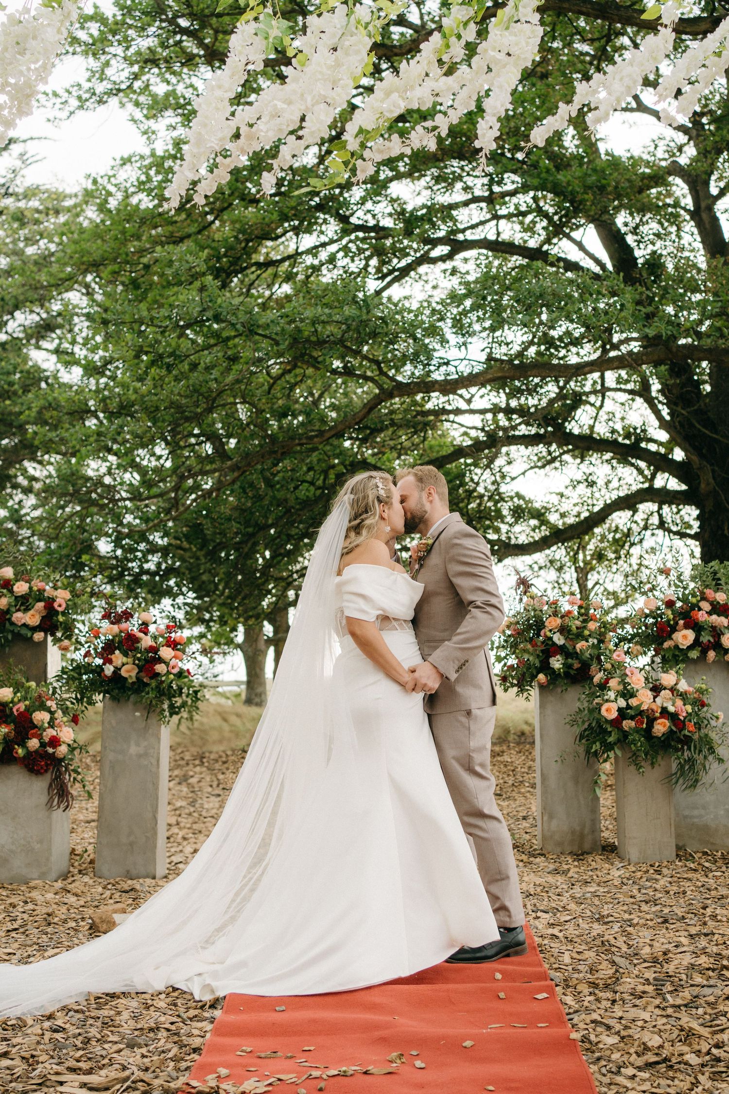 Two figures share a romantic kiss on a red carpet path under flowering trees with pink floral arrangements on pillars.