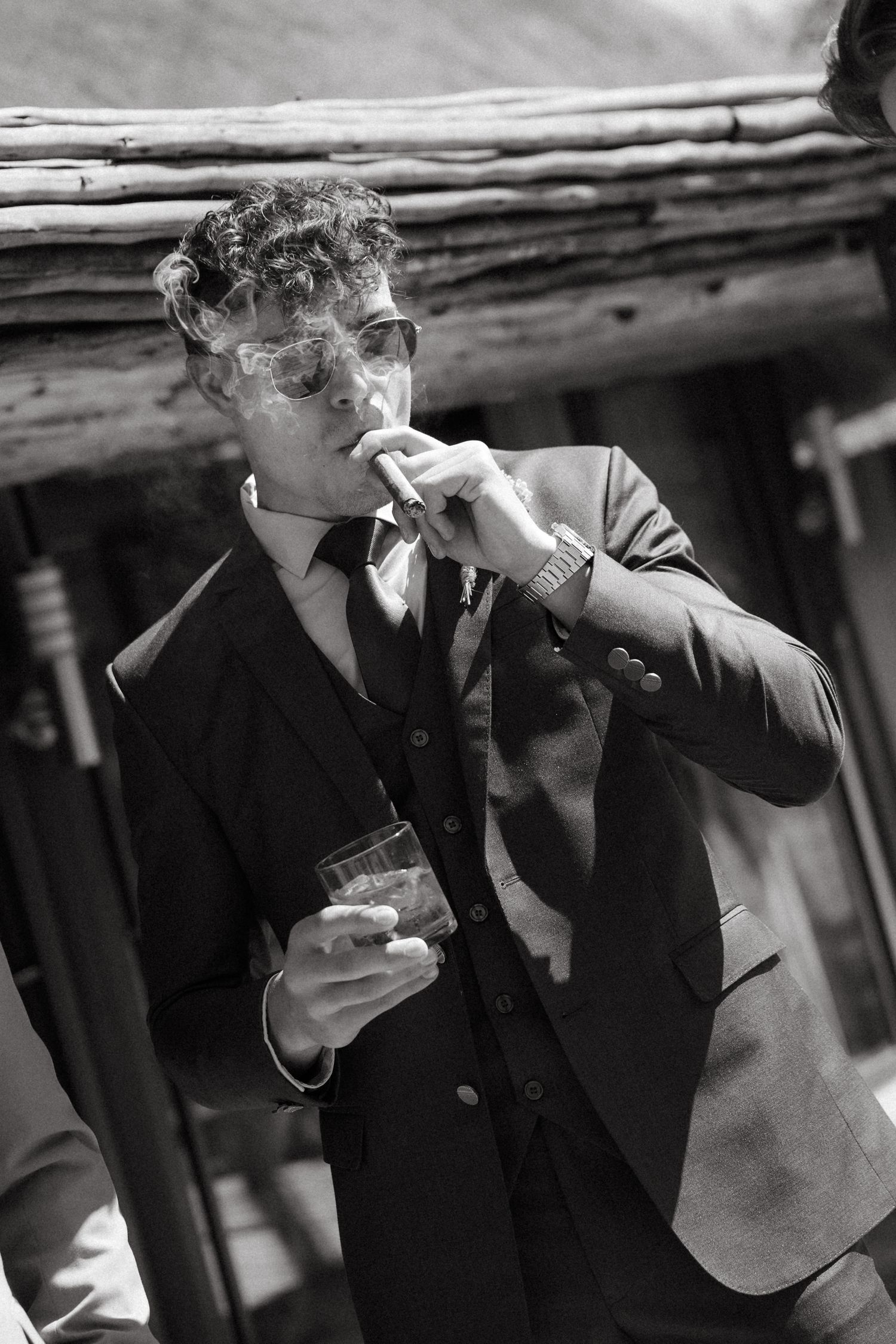 Stylish groom in suit smoking cigar and holding whiskey glass, black and white portrait photography.