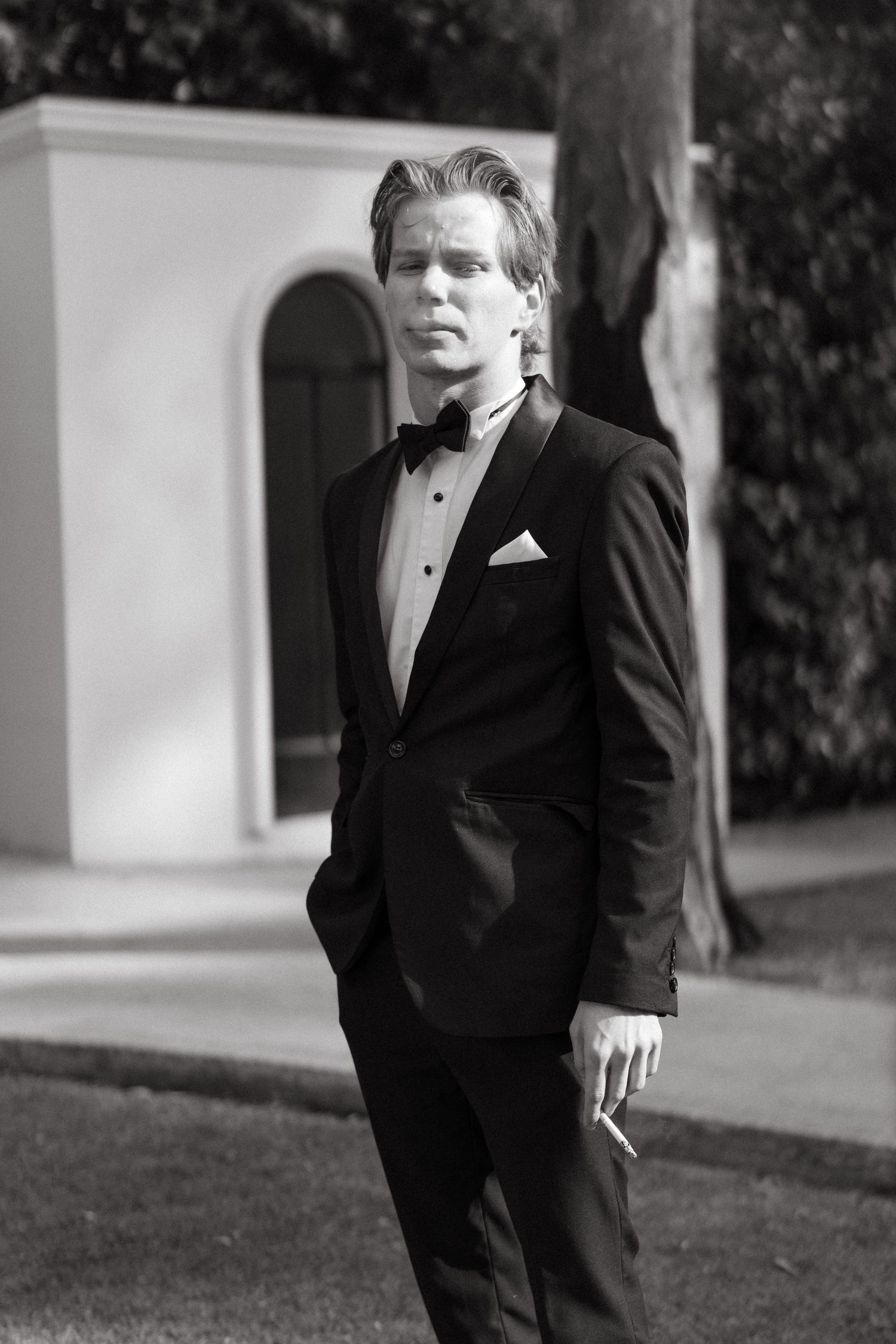 Young man in black tuxedo with bow tie holding cigarette poses outdoors in black and white portrait photo at Lace on Timber.