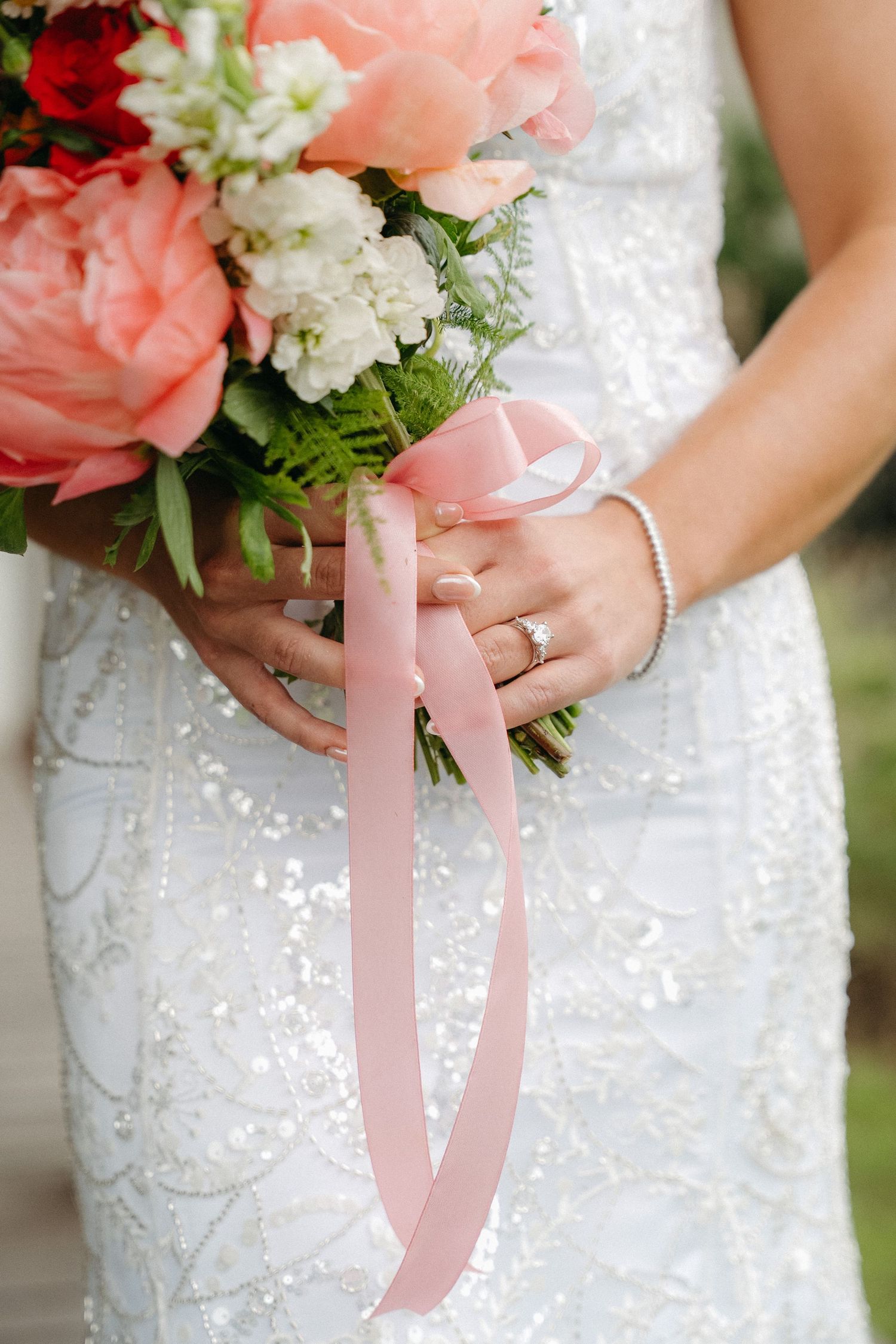 Bride holding a pink and white wedding bouquet with ribbon, wearing a beaded gown and diamond ring.
