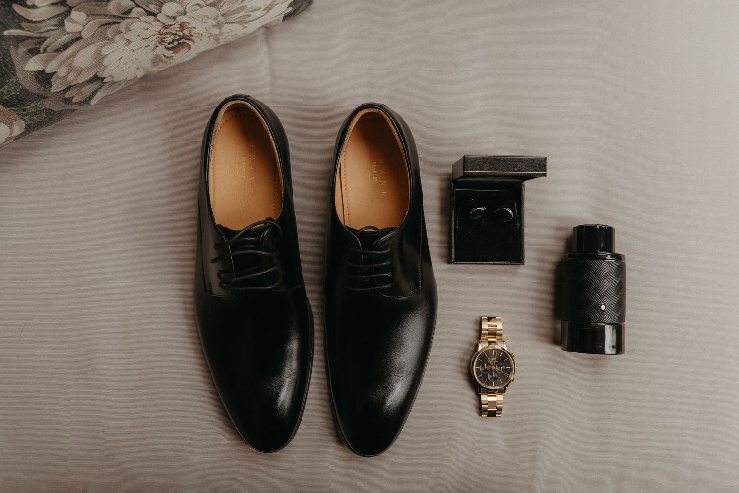 Black dress shoes, gold watch, ring box, and cologne arranged on a gray surface for a groom's wedding day accessories.