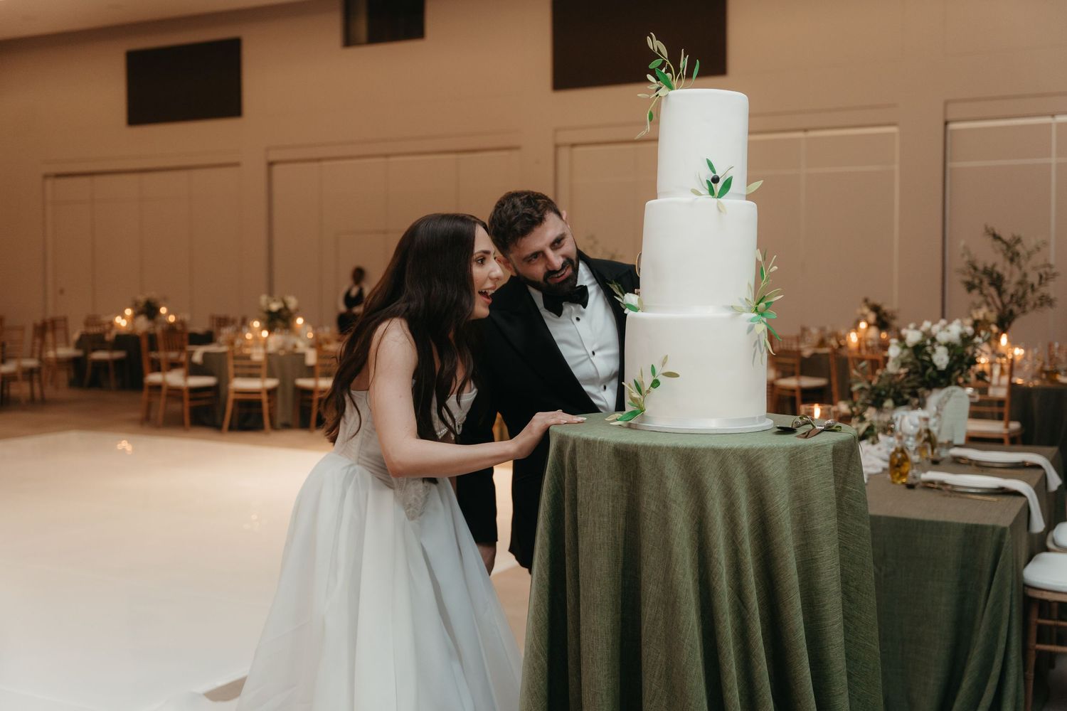 Bride and groom cutting a white four-tier wedding cake decorated with greenery at an elegant reception venue.