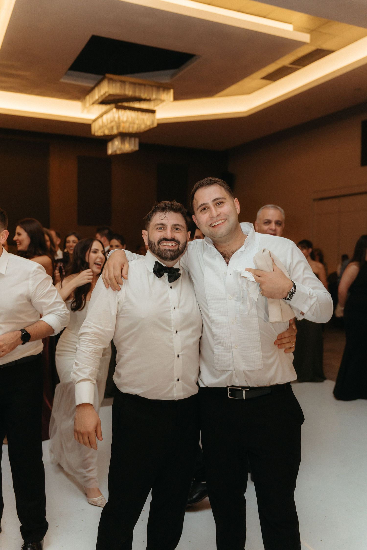 Two men in white shirts smiling and posing together on a dance floor at an elegant wedding reception.