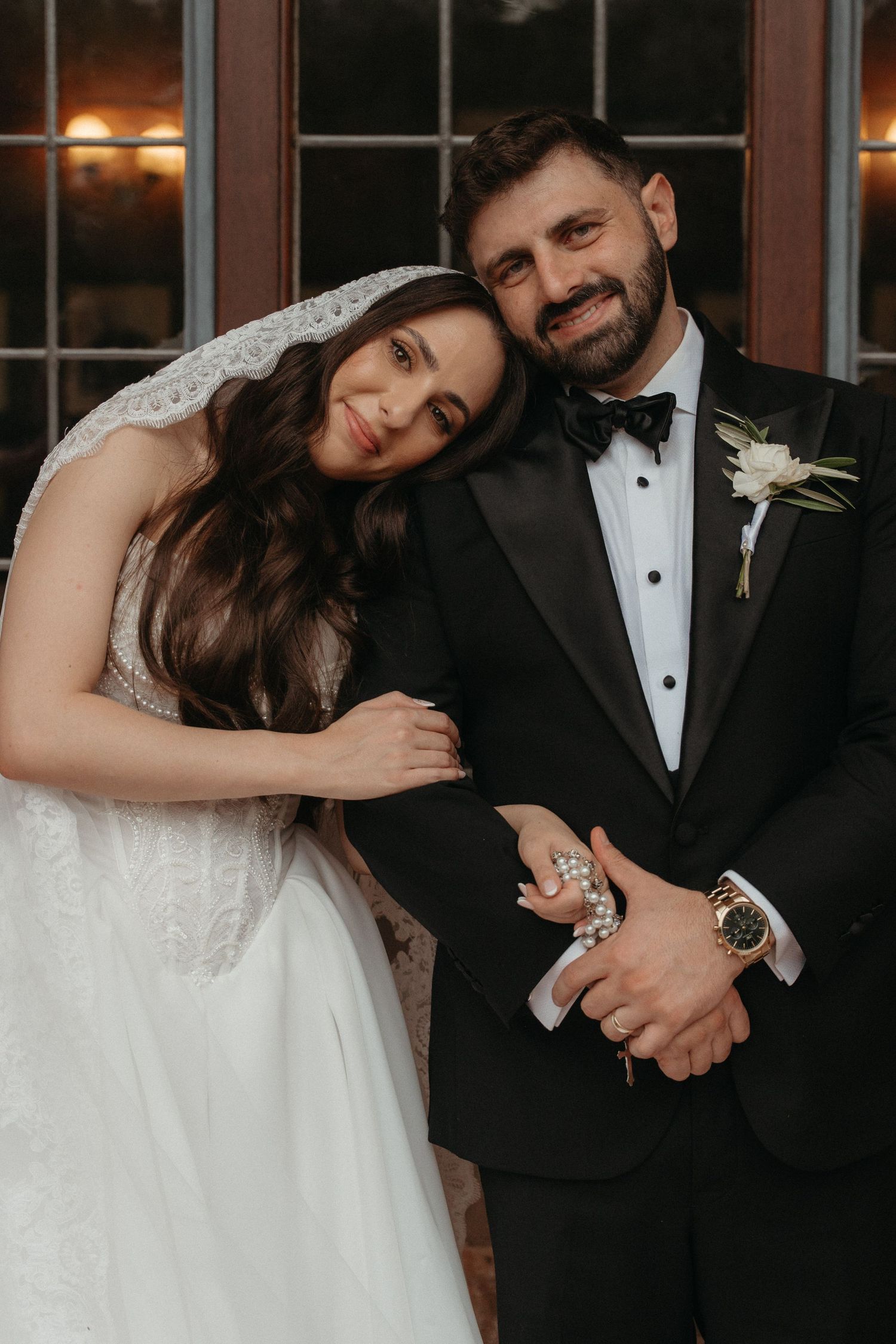 Bride in white lace gown and veil leans on groom in black tuxedo with boutonniere for wedding portrait.