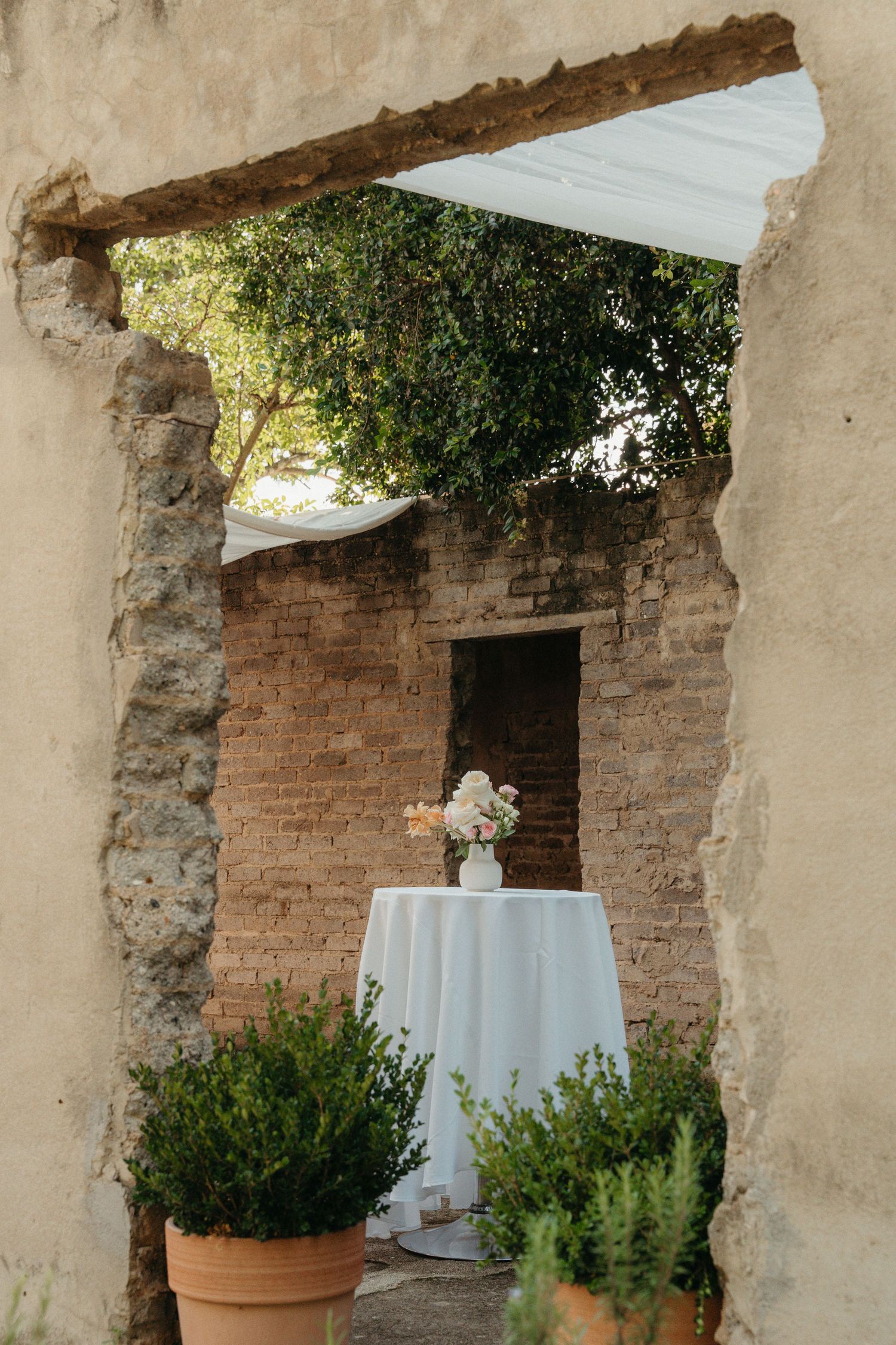 Rustic outdoor wedding cocktail table with white linen and floral arrangement framed by ancient stone archway.