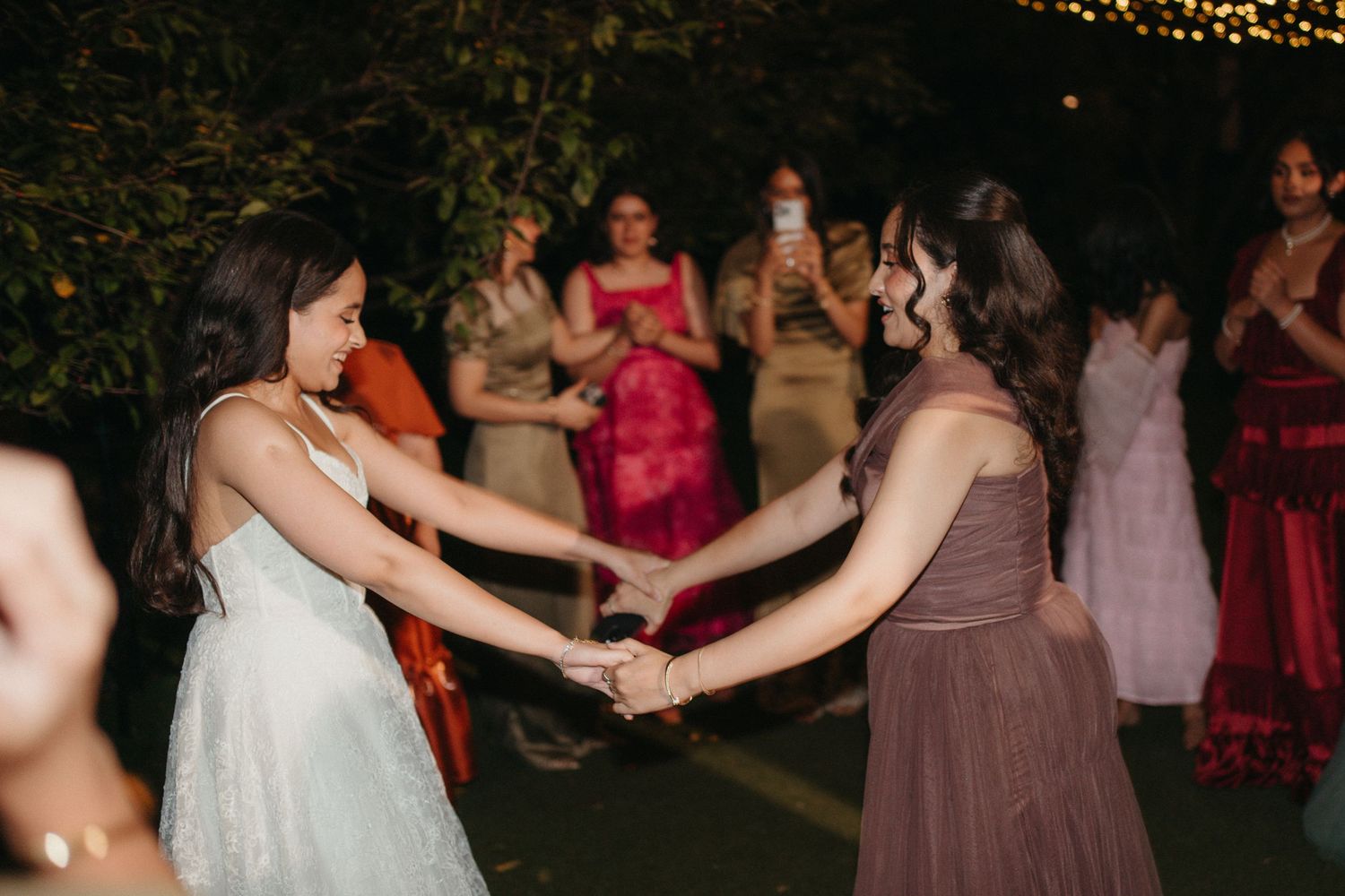 Two women in elegant dresses hold hands and dance joyfully at an outdoor wedding reception with guests watching.