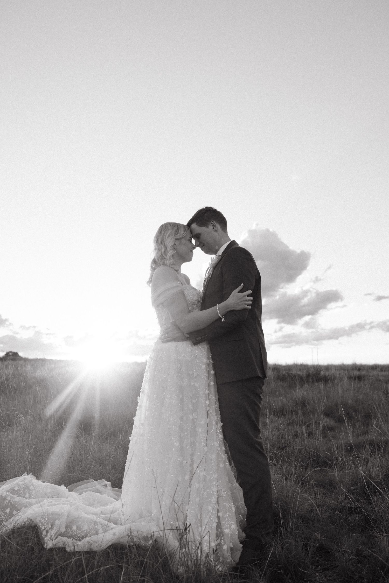Black and white wedding photo of bride and groom embracing at sunset in a field, sun flare behind them.