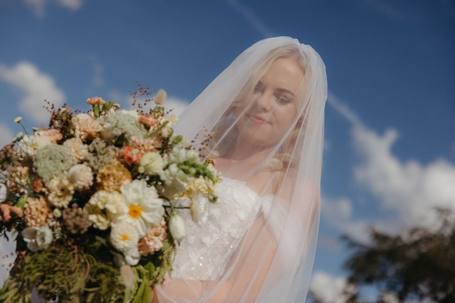 Bride in white veil holding a lush bouquet of white and peach flowers under a blue sky.