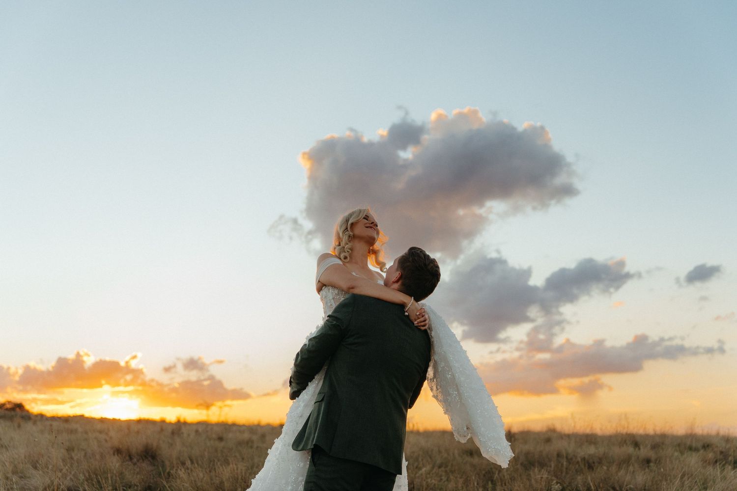 Groom lifting bride at sunset in a golden field, romantic wedding photo with dramatic clouds.