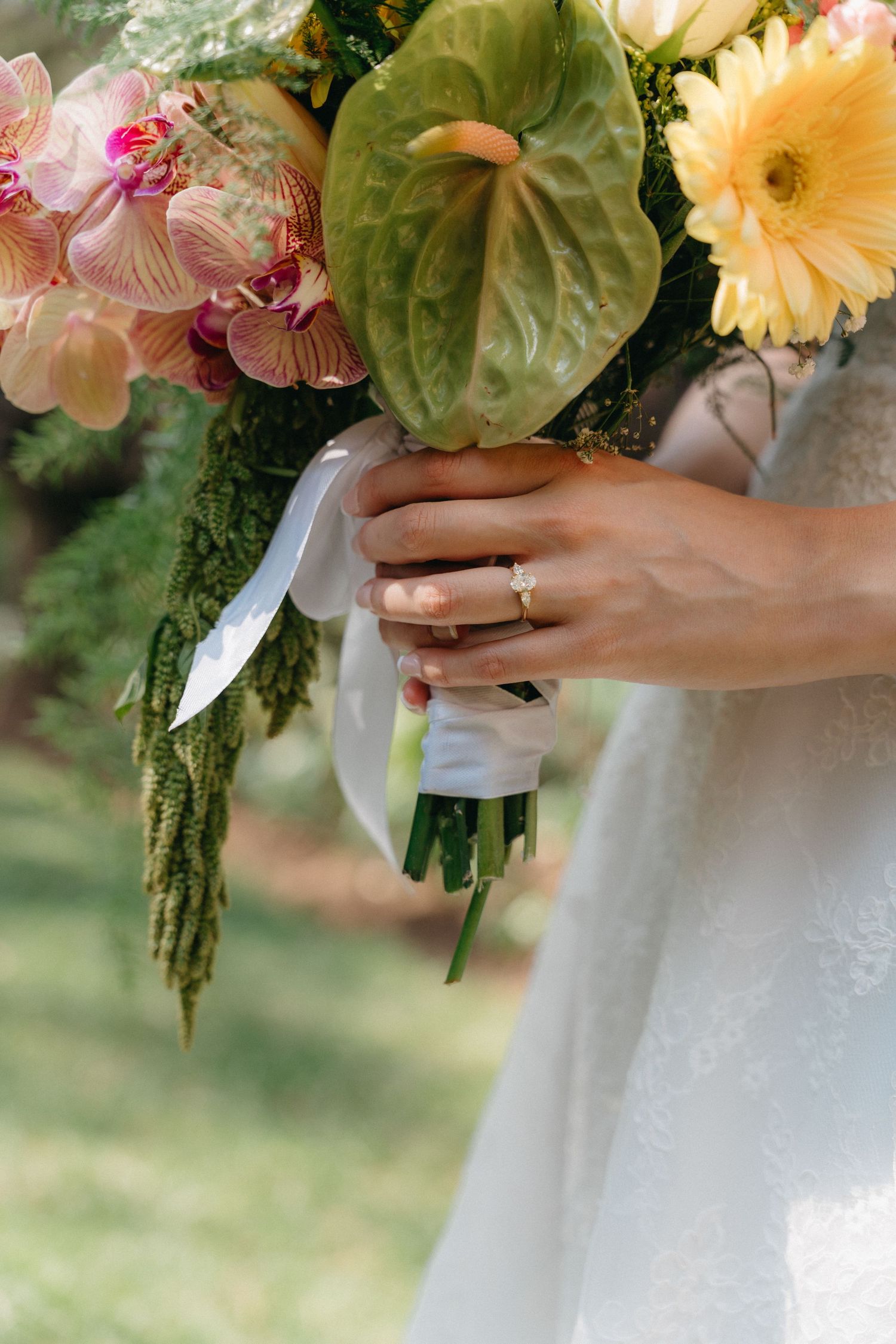 Close-up of bride holding tropical bouquet with anthurium, orchids, and yellow gerbera with ring visible.