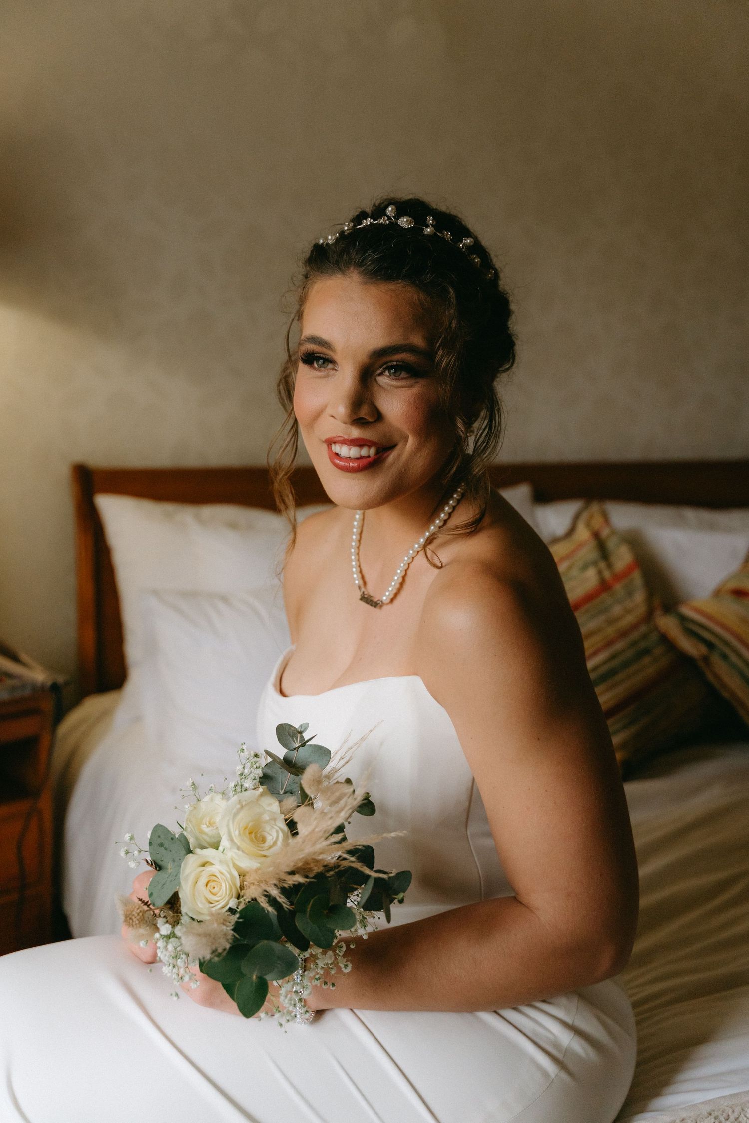 Radiant bride in strapless gown holds white rose bouquet while seated on bed at La La Nathi in Harrismith