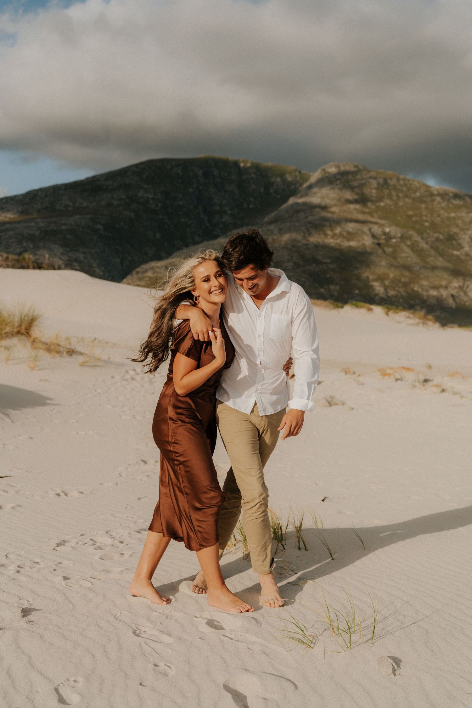 Couple walking barefoot on white sand dunes with mountains in background, woman in brown satin dress and man in white shirt.
