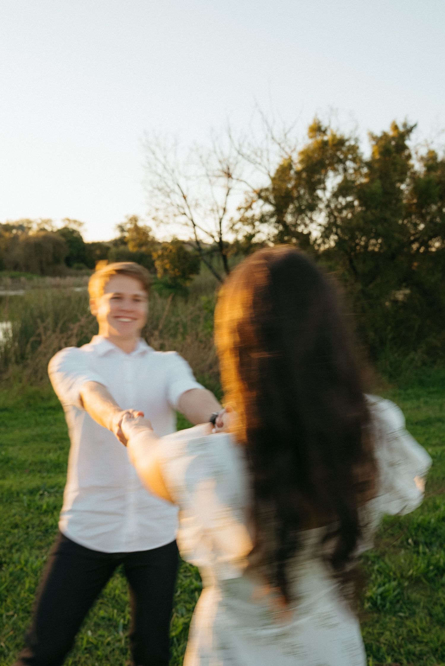 Two people hold hands and spin together in a sunlit grassy field during golden hour.