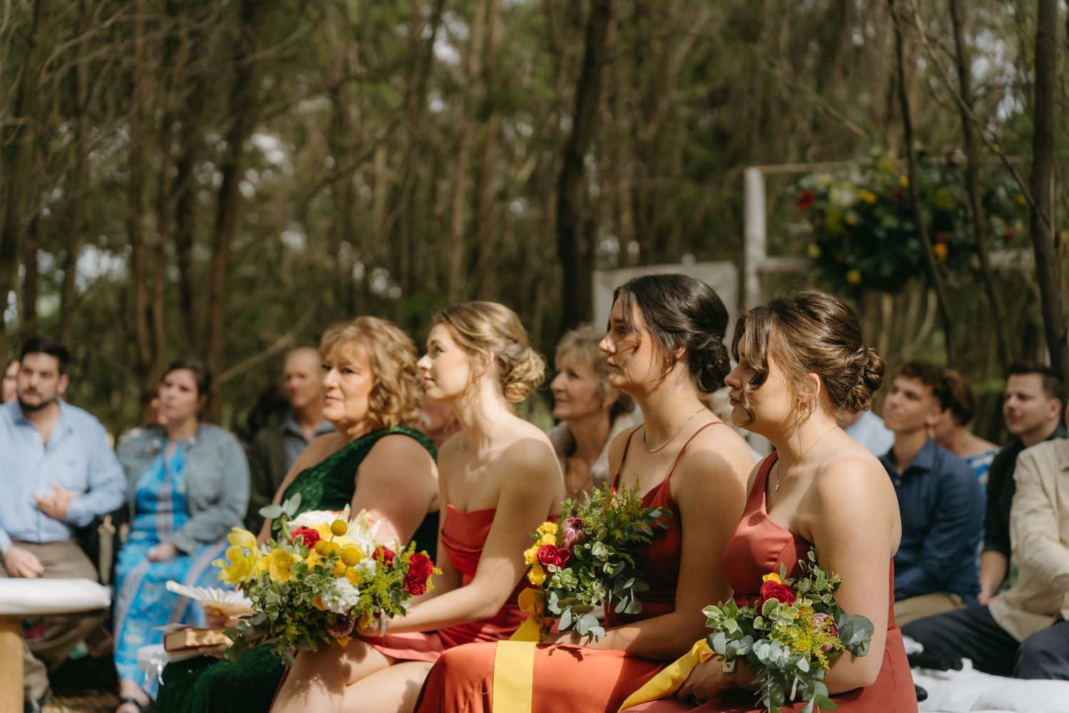 Bridesmaids in red dresses hold colorful wildflower bouquets during an outdoor forest wedding ceremony.
