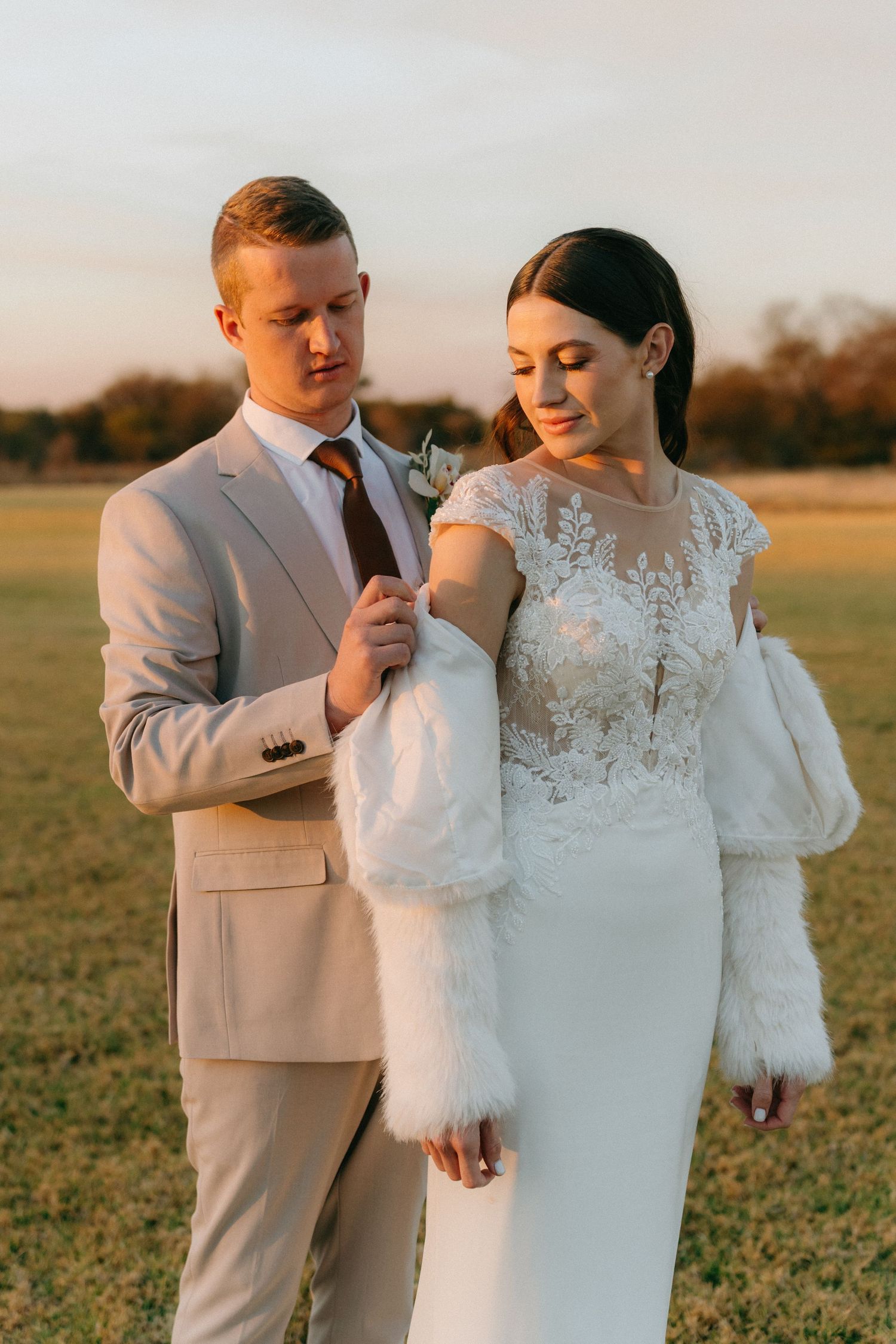 Elegant couple in light-colored wedding attire pose together in a sunlit field during golden hour.