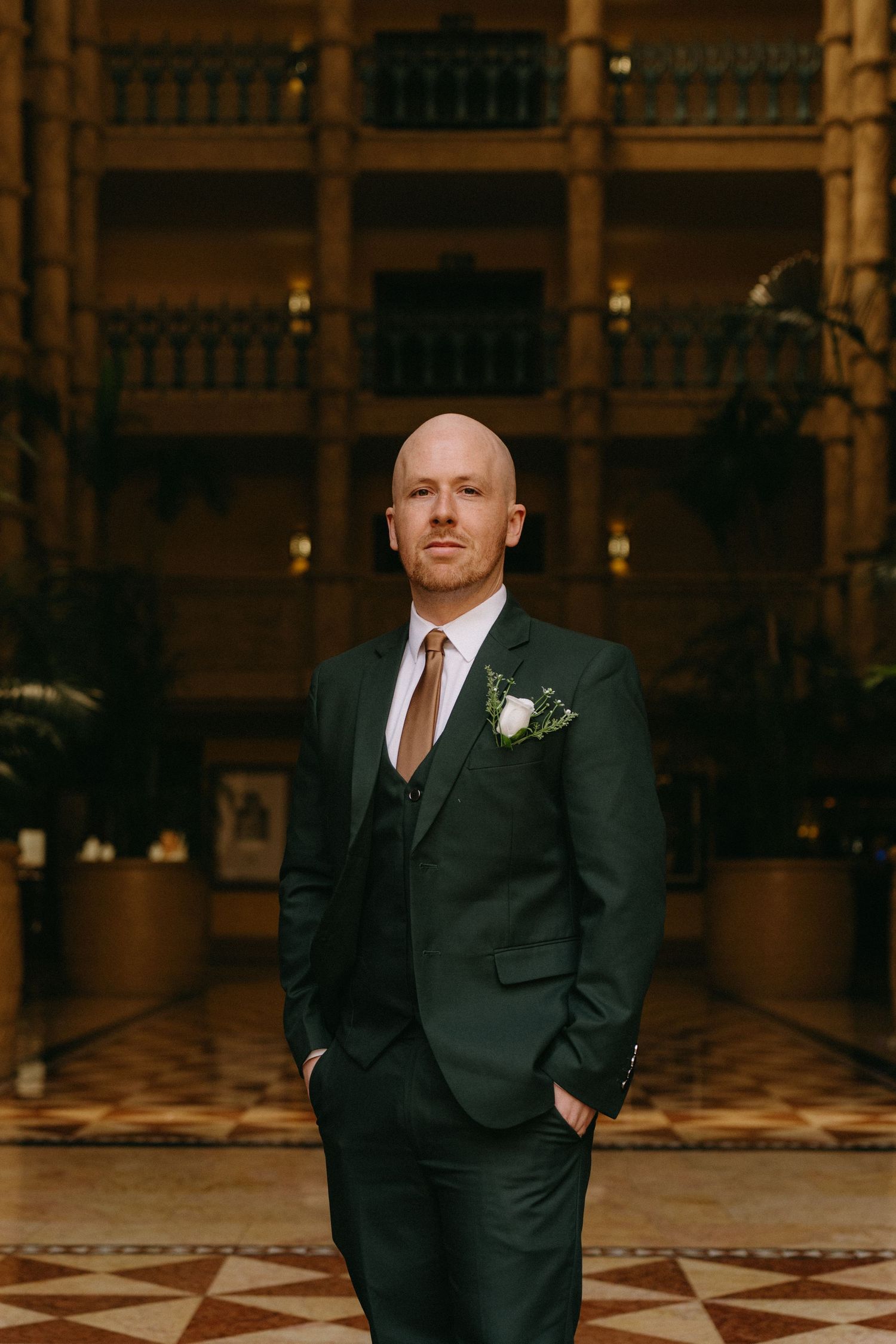 A person in a dark formal suit with a boutonniere poses in an elegant hotel lobby with ornate tile flooring.
