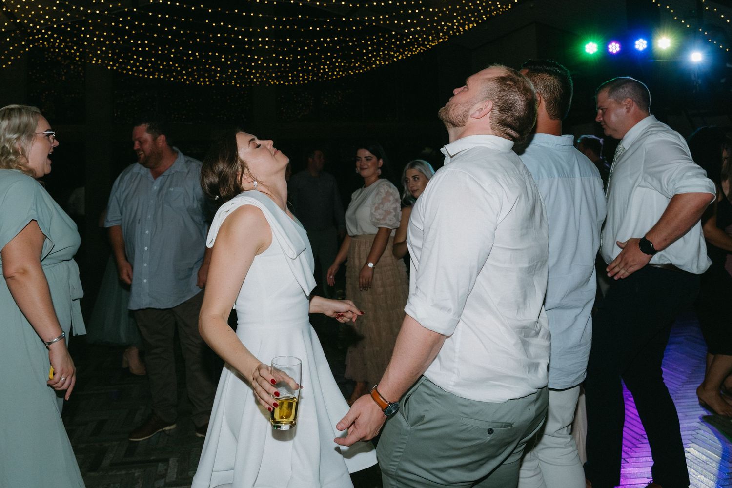 People dancing and mingling at an evening reception under twinkling string lights and colorful stage lighting.