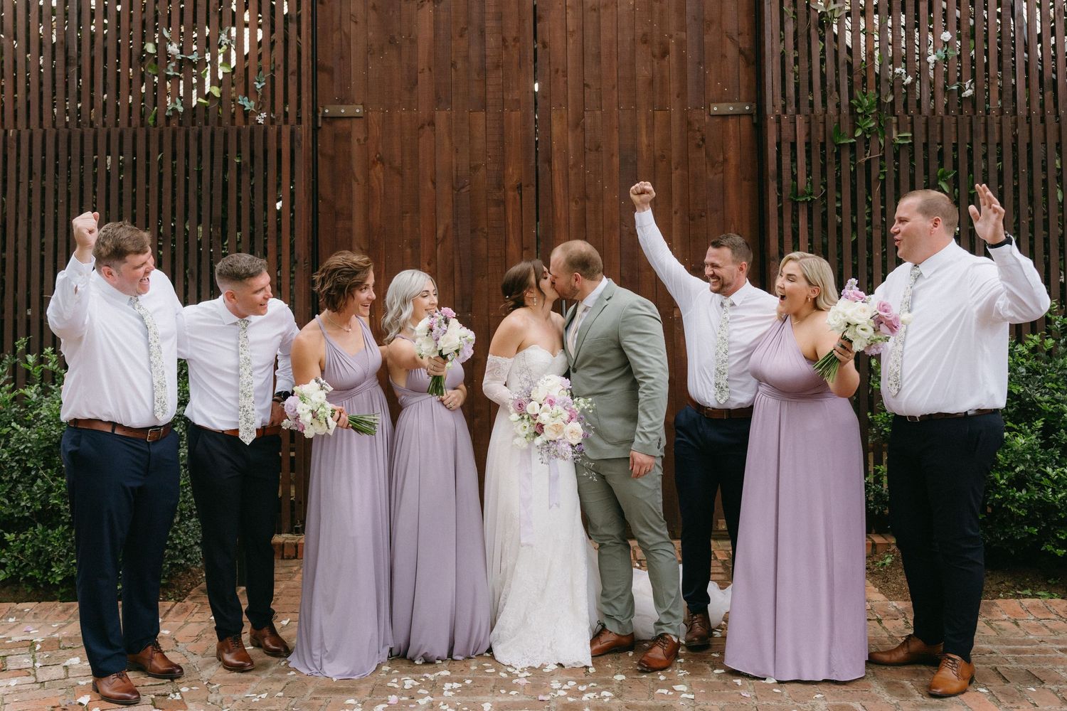 Wedding party celebrates joyfully against wooden wall backdrop with bridesmaids in lavender dresses.