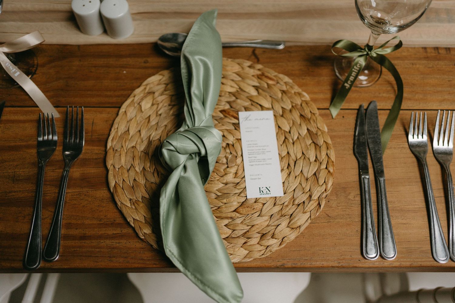 Table setting with sage green napkin, woven placemat, and elegant silverware on wooden surface.